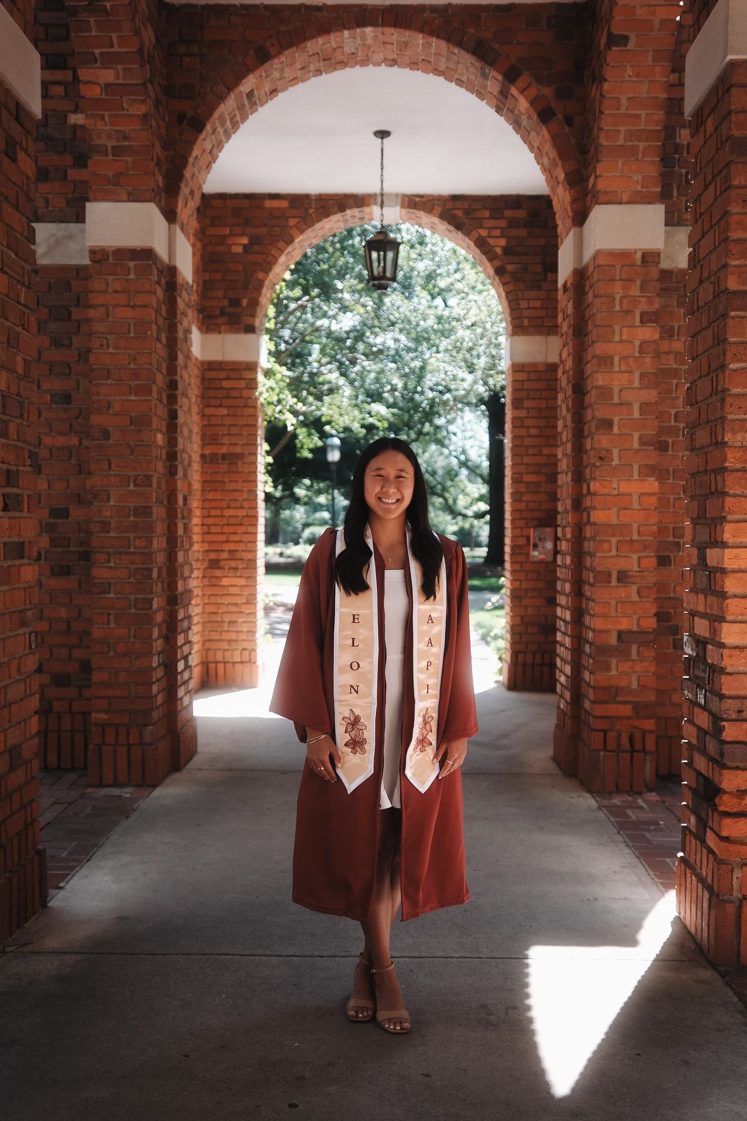 Elon University graduate in maroon gown and stole smiling under brick archway on campus.