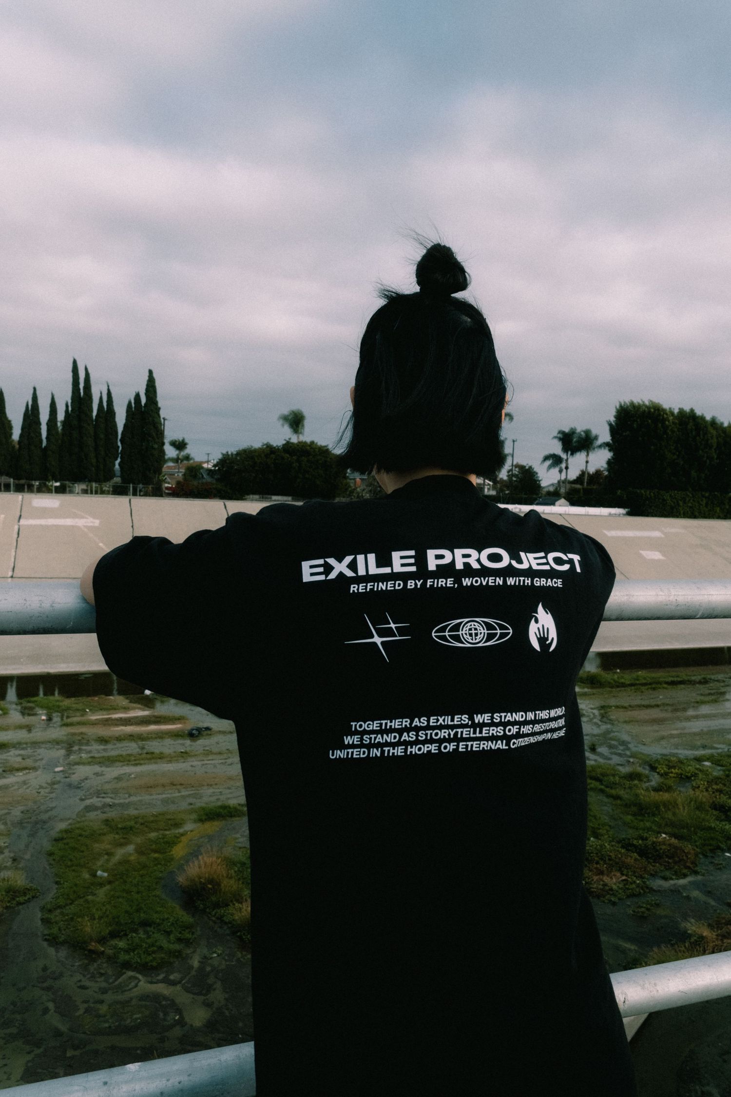 Person wearing black Exile Project long sleeve shirt with graphic print, standing outdoors near a guardrail.
