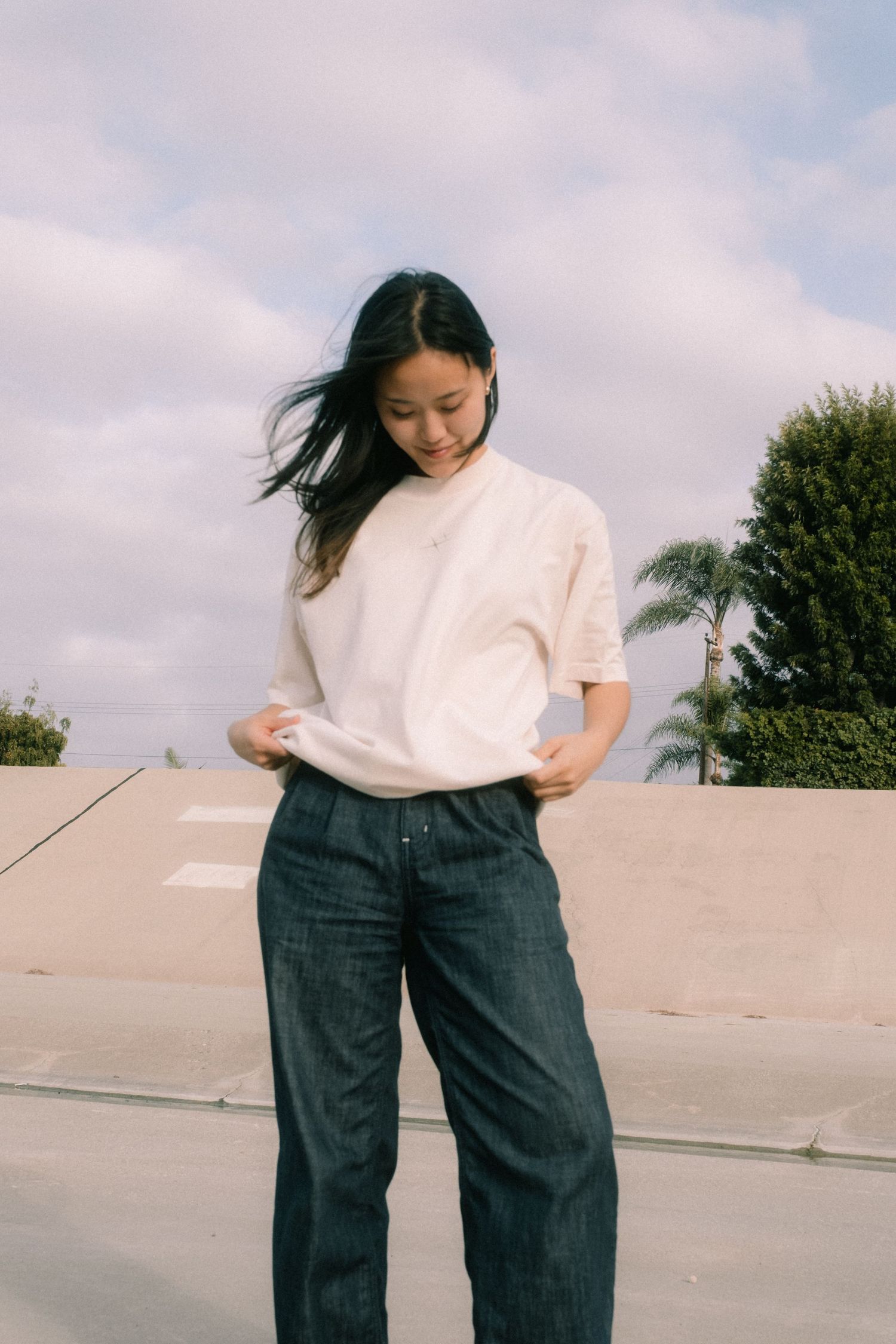 Young woman in white t-shirt and dark wide-leg jeans standing outdoors with wind in her hair.