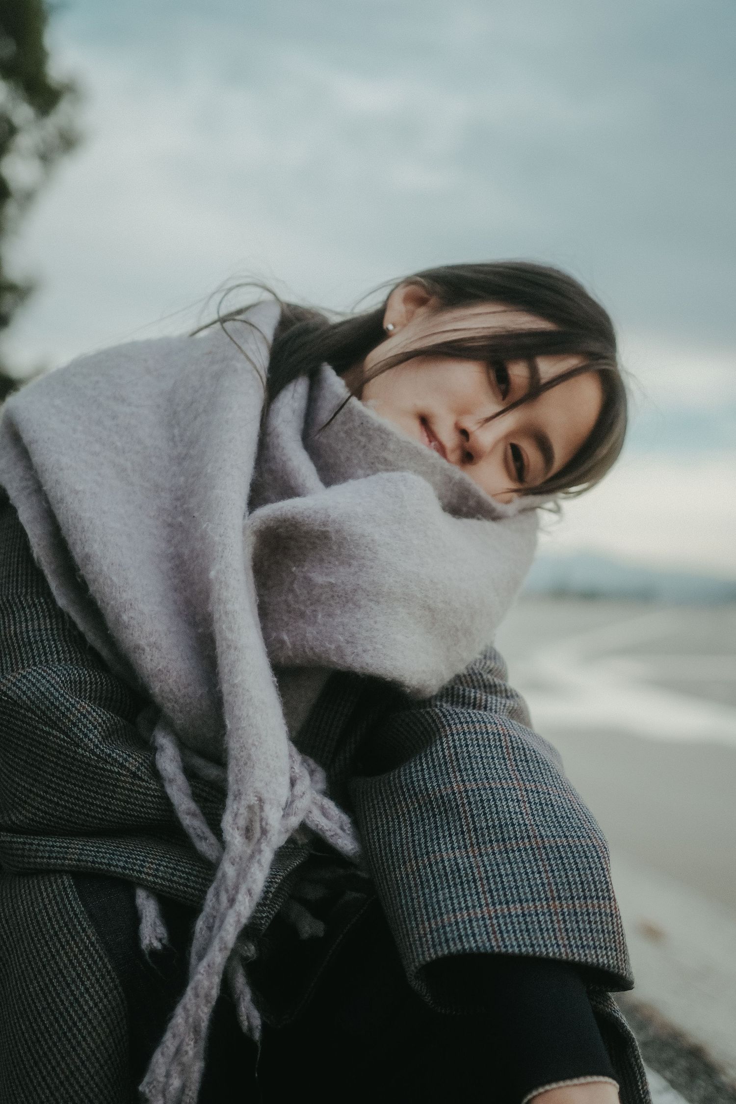 Young woman in a gray plaid coat and oversized scarf sits outdoors by a beach with eyes closed on a cloudy day.