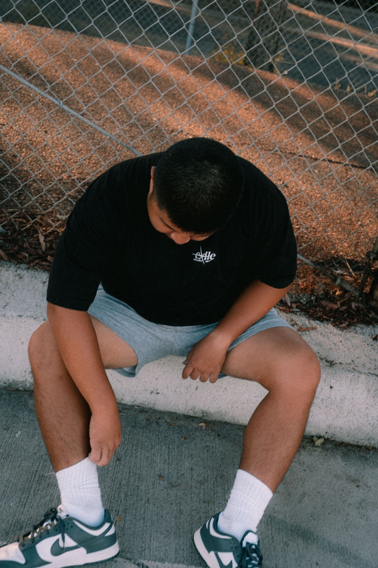 Young man in black tee, gray shorts, and teal Nike Dunks sitting on a curb by a chain-link fence.