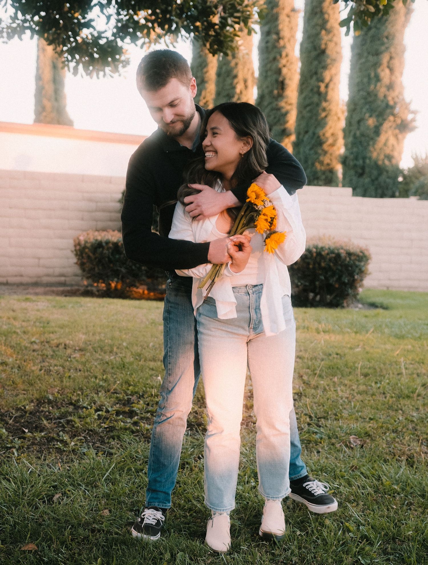 A loving couple hugs outdoors at golden hour, woman holding a bouquet of sunflowers.