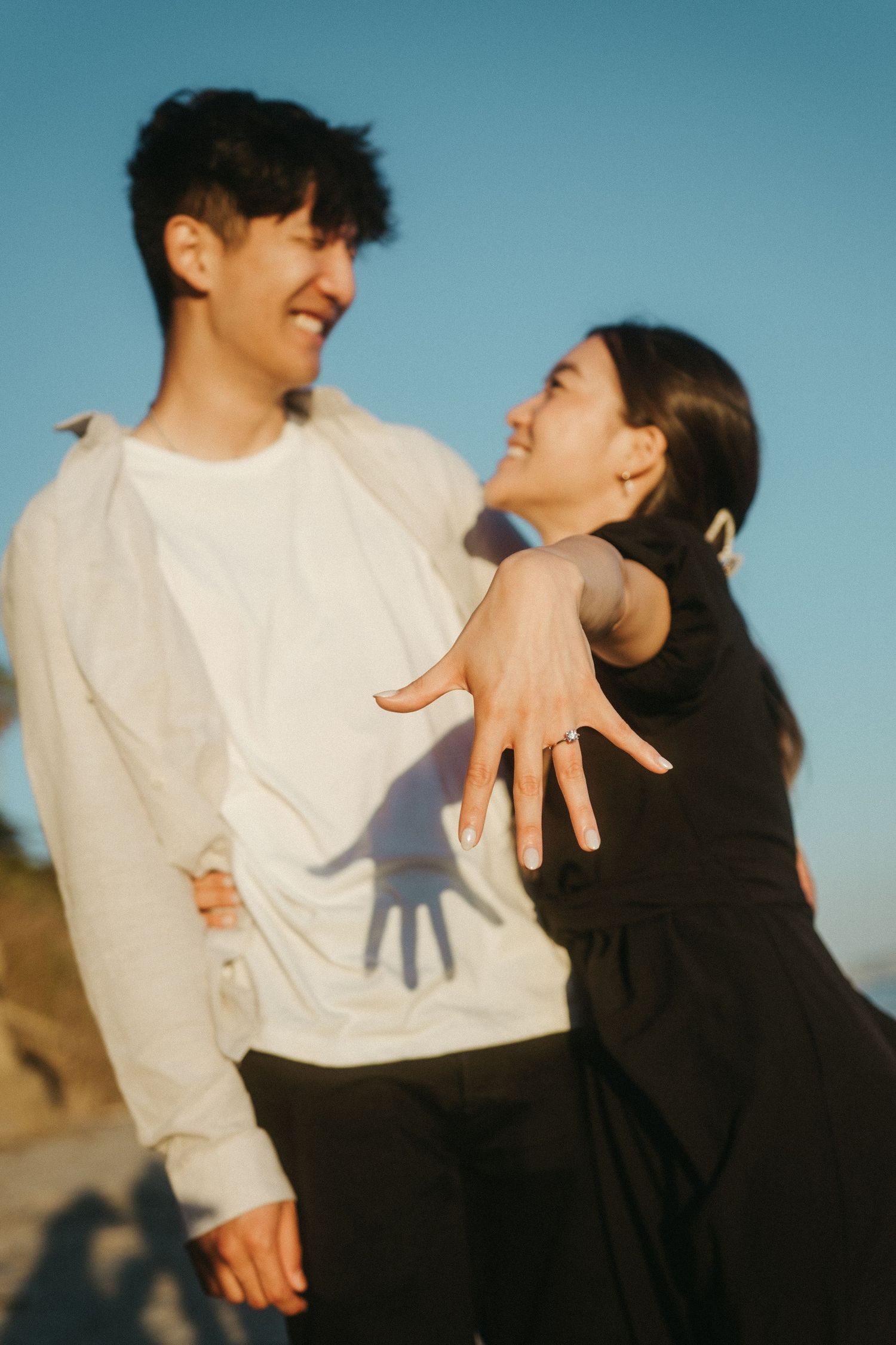 Joyful engaged couple on a sunny beach as woman shows off her diamond engagement ring.
