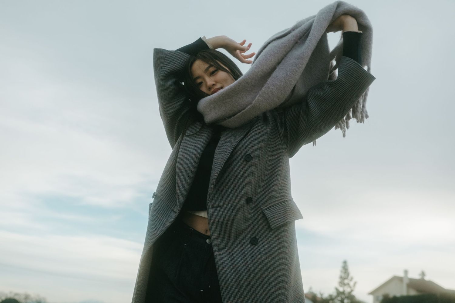 Woman in oversized gray coat and scarf posing outdoors against a cloudy sky.