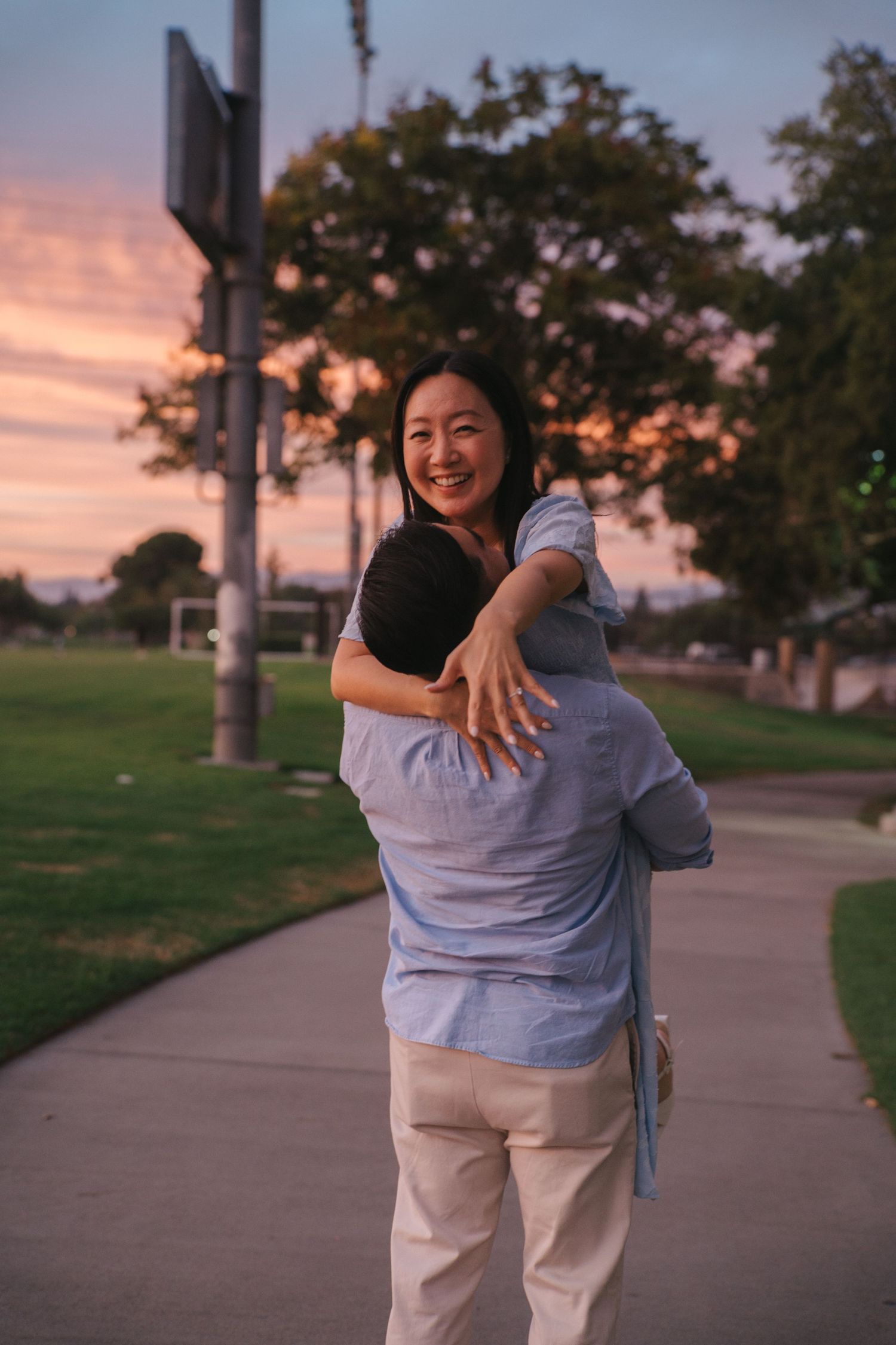 Joyful Asian woman smiling while being lifted in hug by partner on park path at sunset.