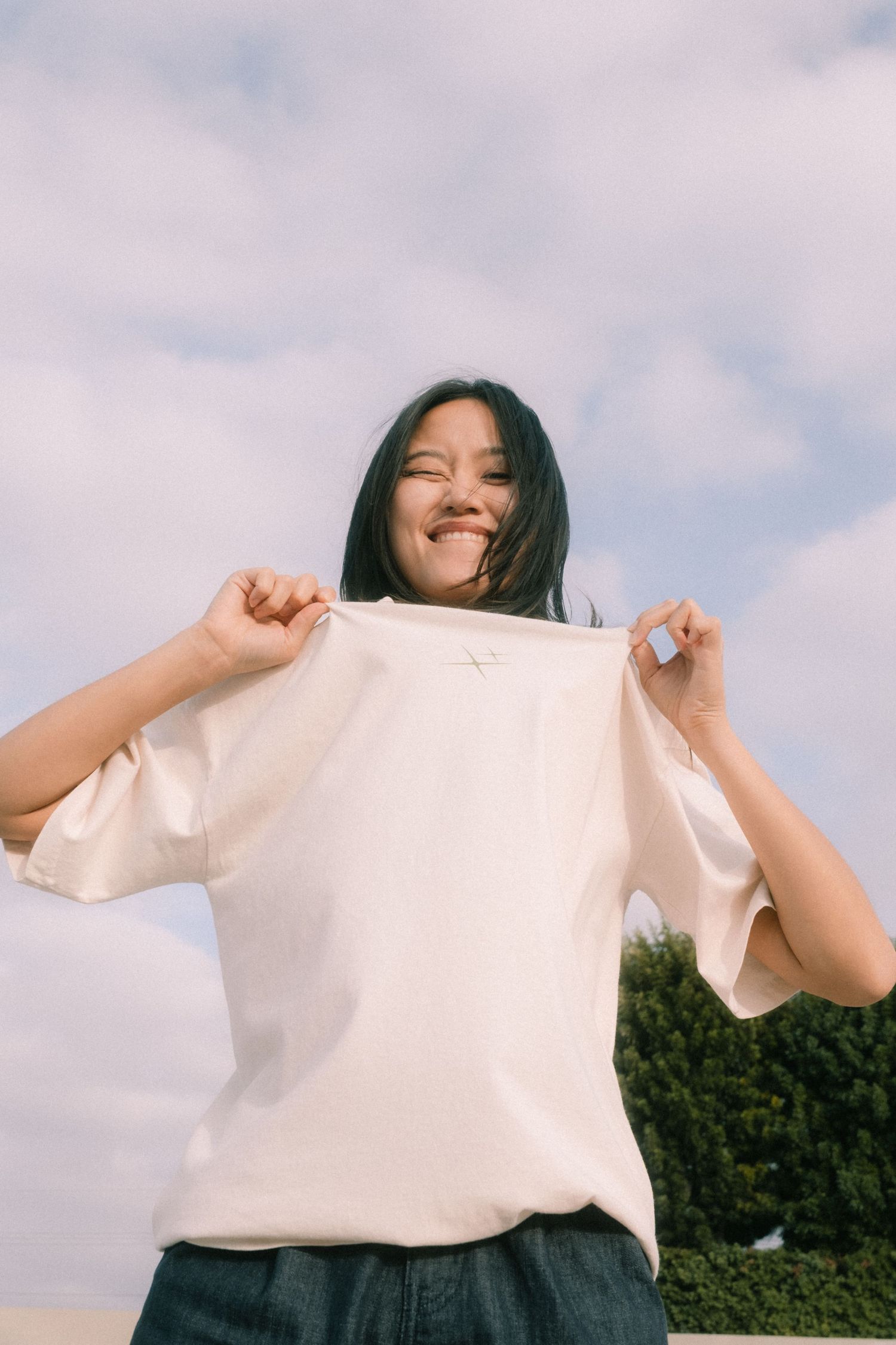 Smiling woman holding up white oversized t-shirt with small cross graphic against cloudy sky.