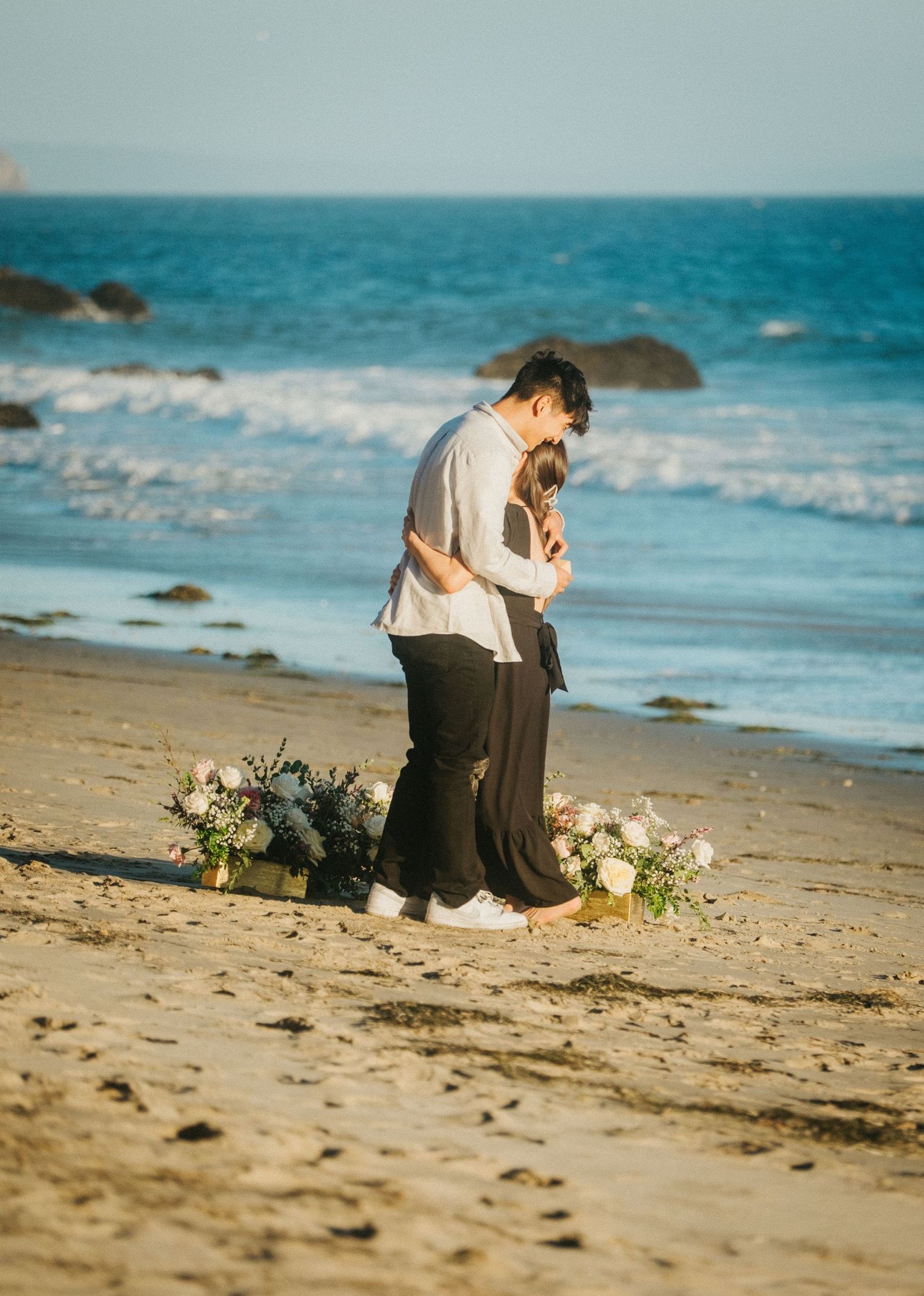 Couple embracing tenderly on a beach after a marriage proposal with flowers on the sand.