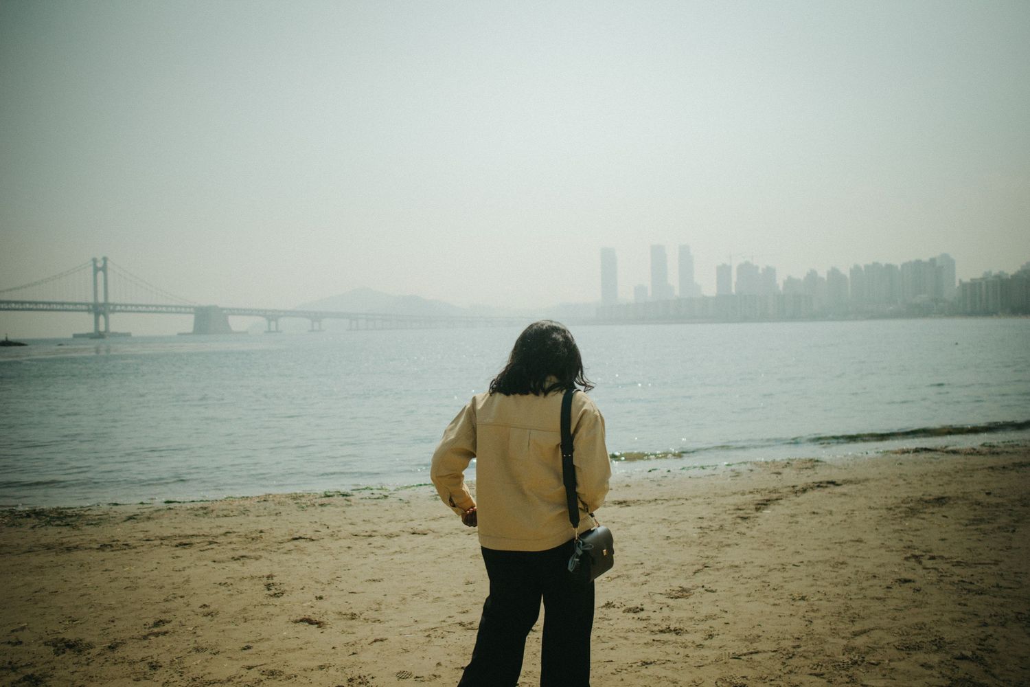 Woman in yellow jacket standing on a sandy beach overlooking a city skyline in hazy weather.
