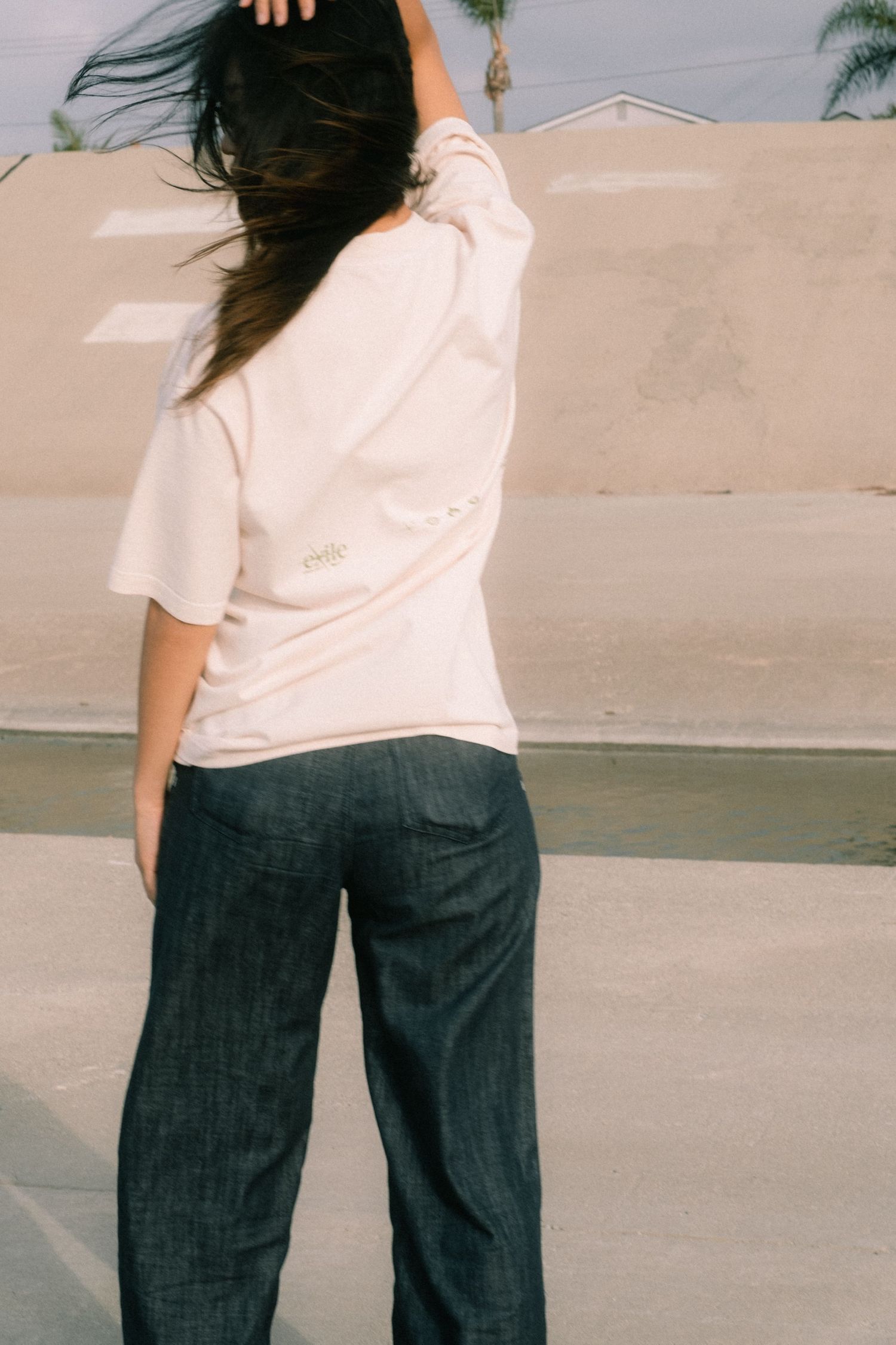 Woman in cream Exile Project t-shirt and dark jeans standing outdoors with wind-blown hair.