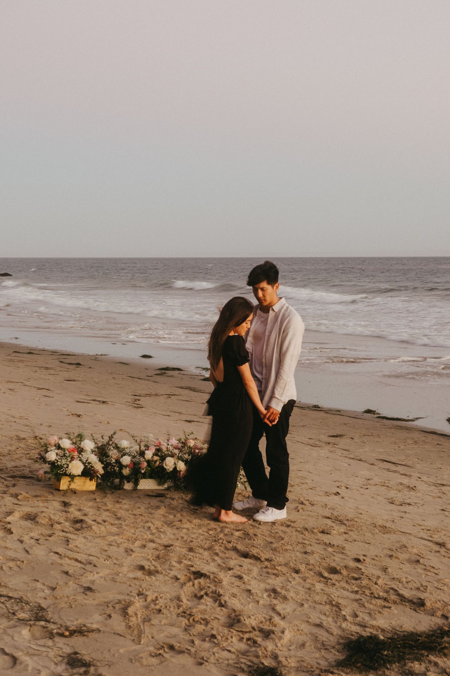 Couple standing together on sandy beach at sunset with floral arrangement nearby.