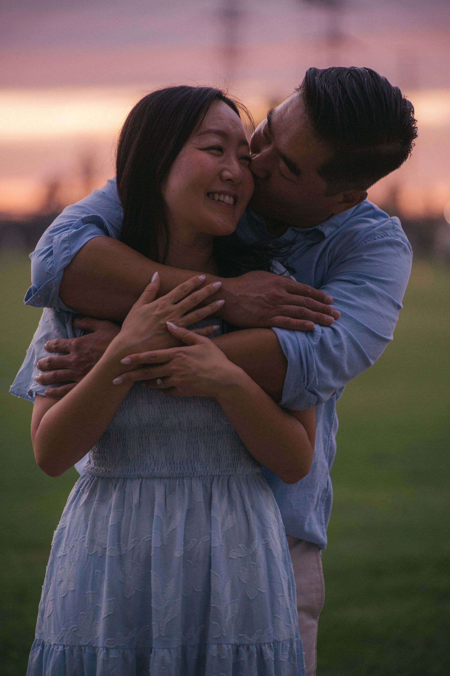 Happy Asian couple embracing at sunset, man kissing womans cheek as she smiles and shows ring.