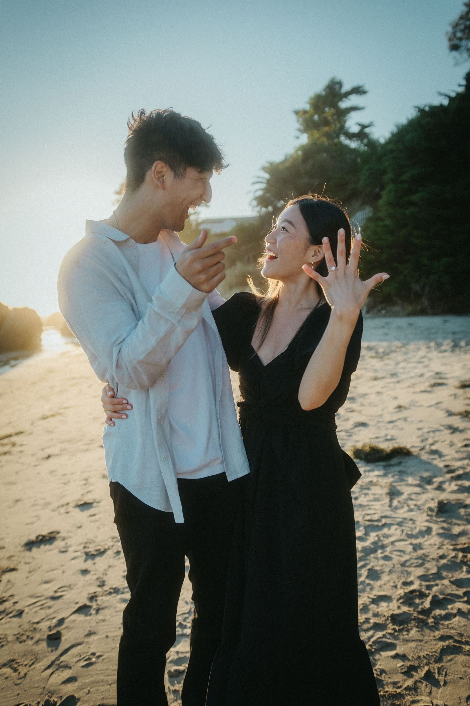 Excited couple on a beach at sunset as woman proudly displays her new engagement ring.