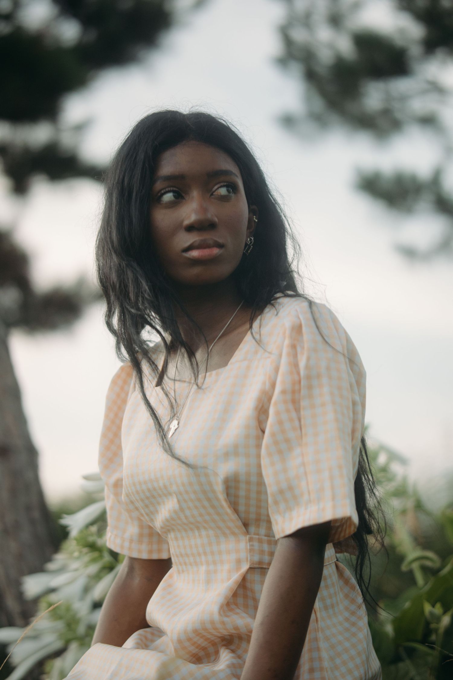 Young Black woman in gingham dress gazing upward among trees and white flowers.