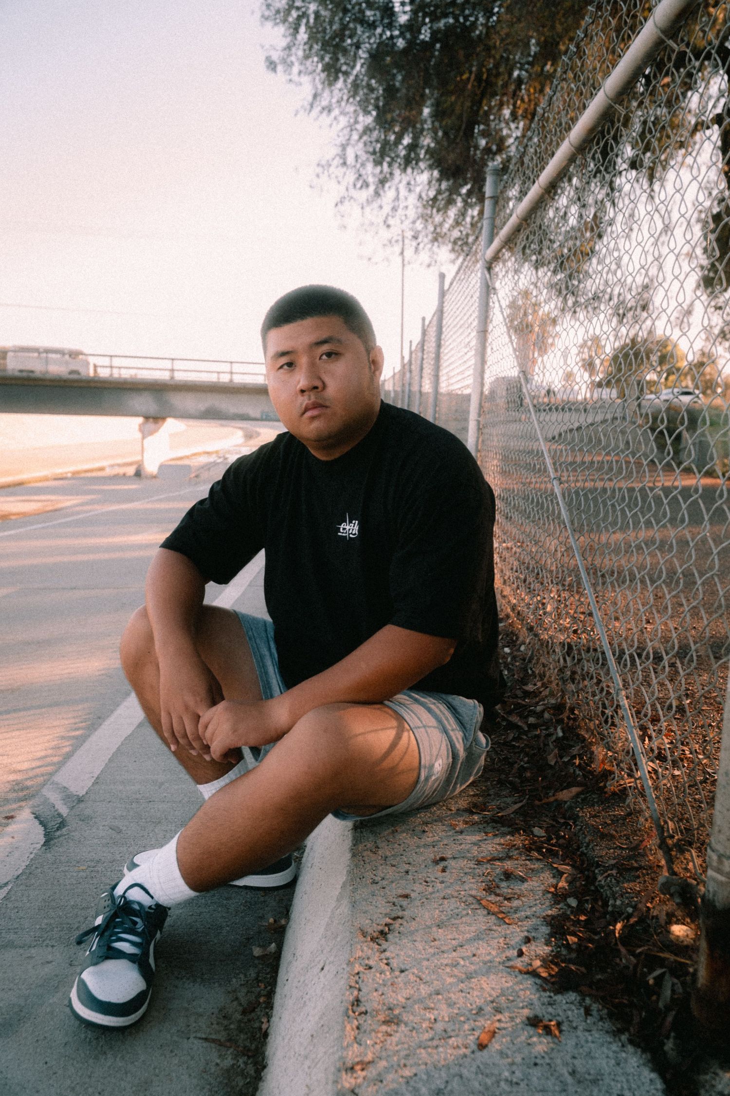 Young man in black tee and shorts sitting by chain-link fence at sunset.