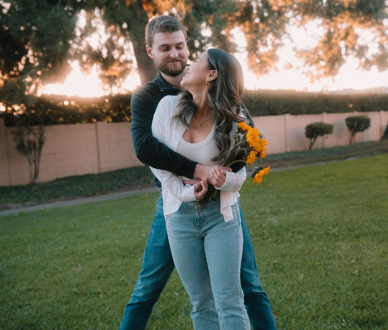 A happy couple dances together in a park at sunset, woman holding bright yellow flowers.