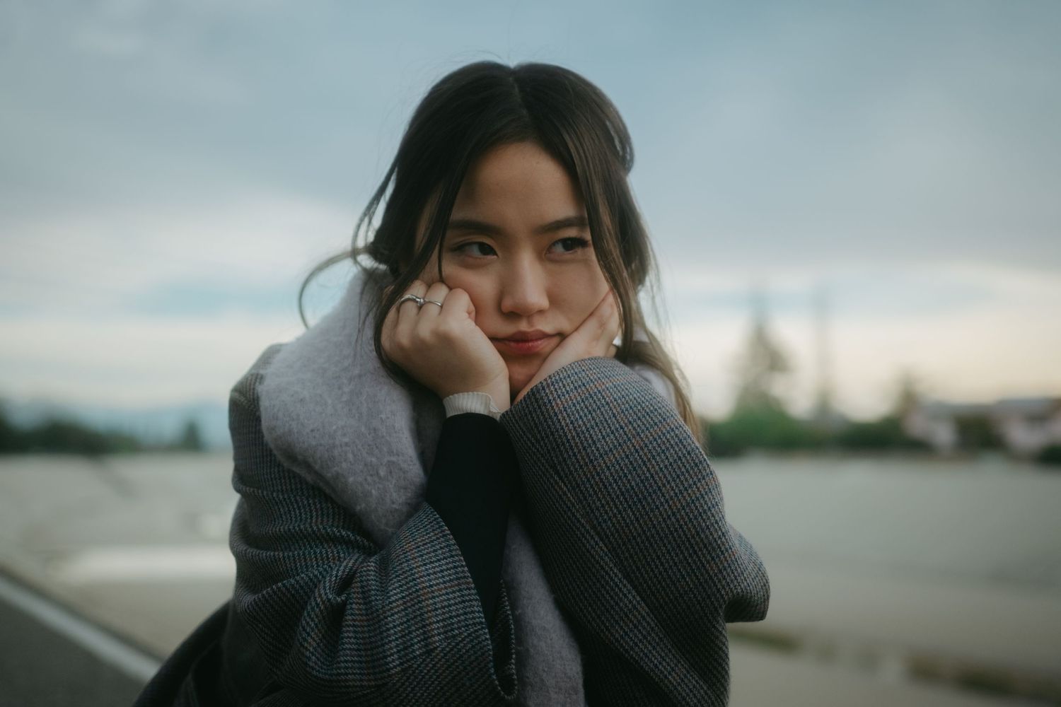 Young Asian woman in gray coat posing thoughtfully by waterfront at dusk, hands framing her face.