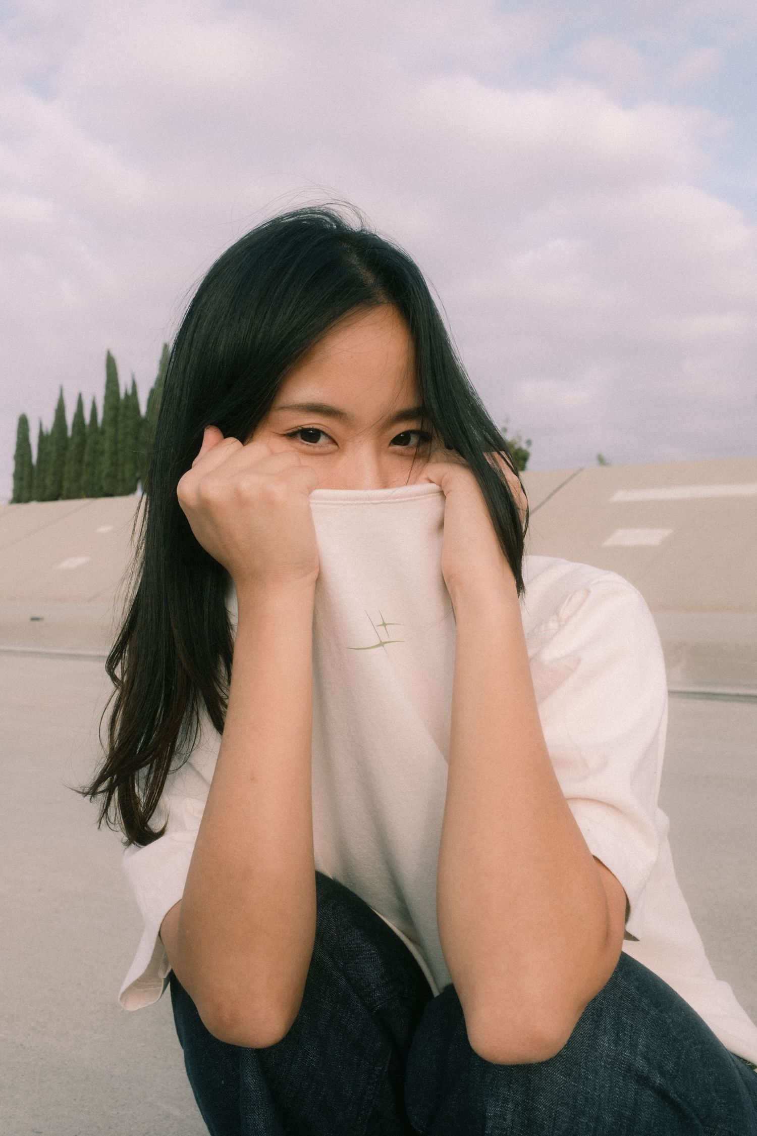 Young Asian woman sitting outdoors, playfully hiding her face behind her white top, with cloudy sky and trees behind her.