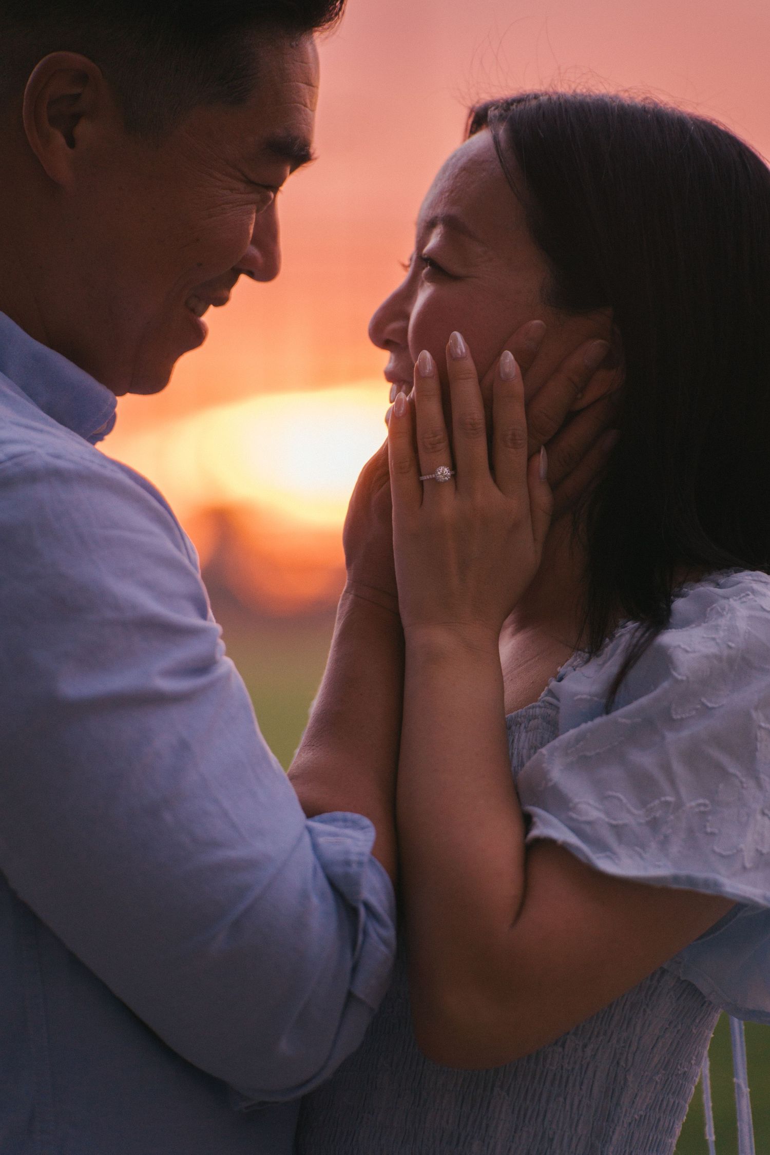 Romantic couple sharing a tender moment at sunset, man gently cupping womans face as she smiles wearing an engagement ring.