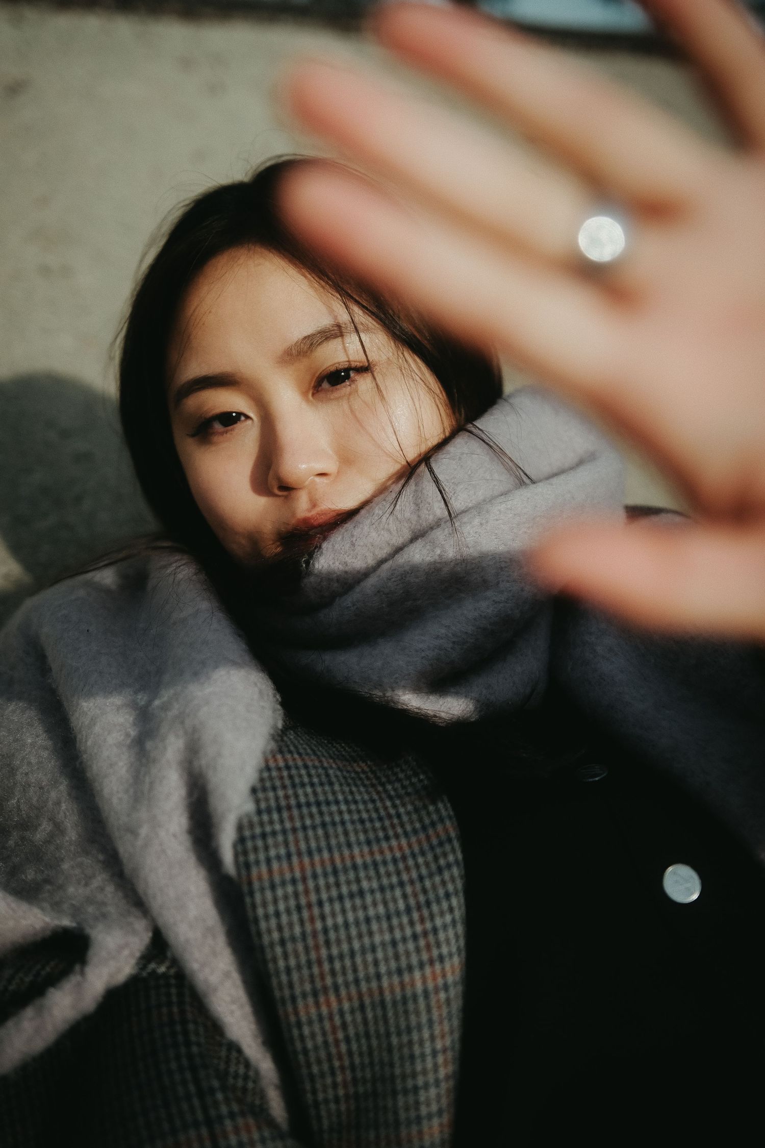 Young Asian woman in gray scarf and coat reaching hand toward camera in artistic portrait photo.