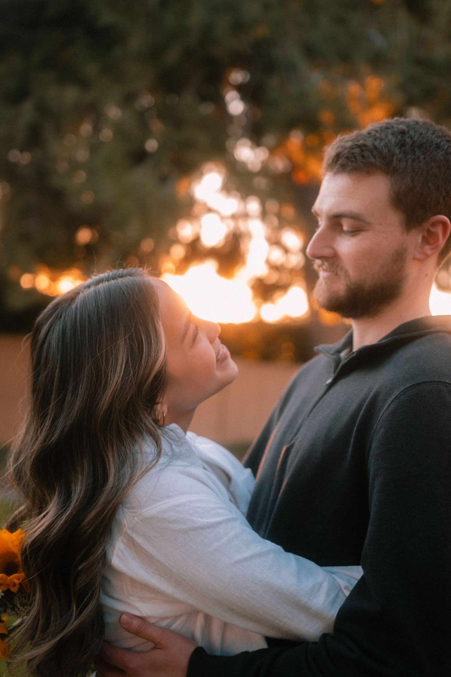 Couple embracing at golden hour sunset, woman holding sunflowers, romantic outdoor engagement photo.