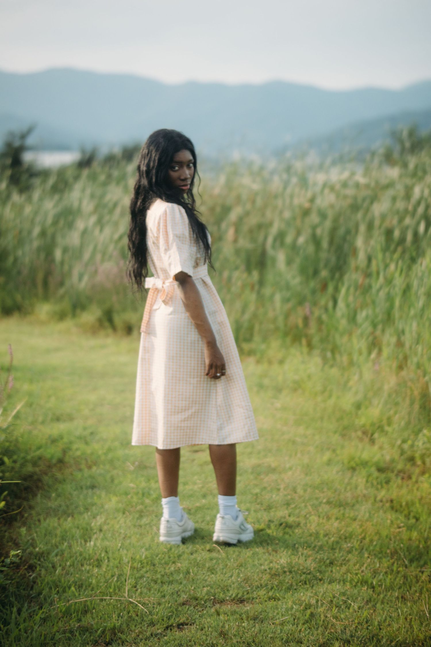 Young Black woman in white midi dress stands on grassy path with tall reeds behind.