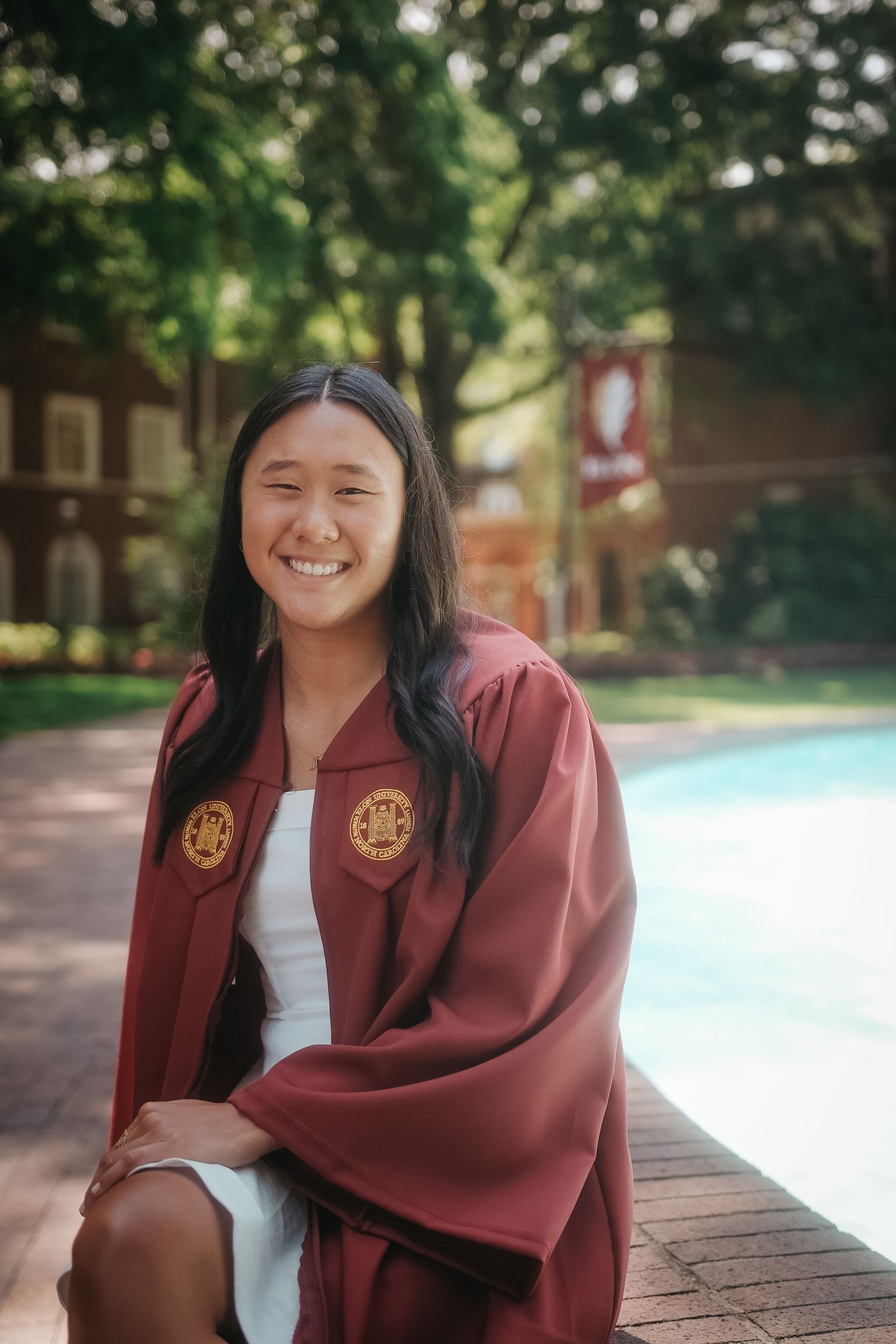 Smiling Elon University graduate in maroon gown sitting beside campus fountain on brick ledge.