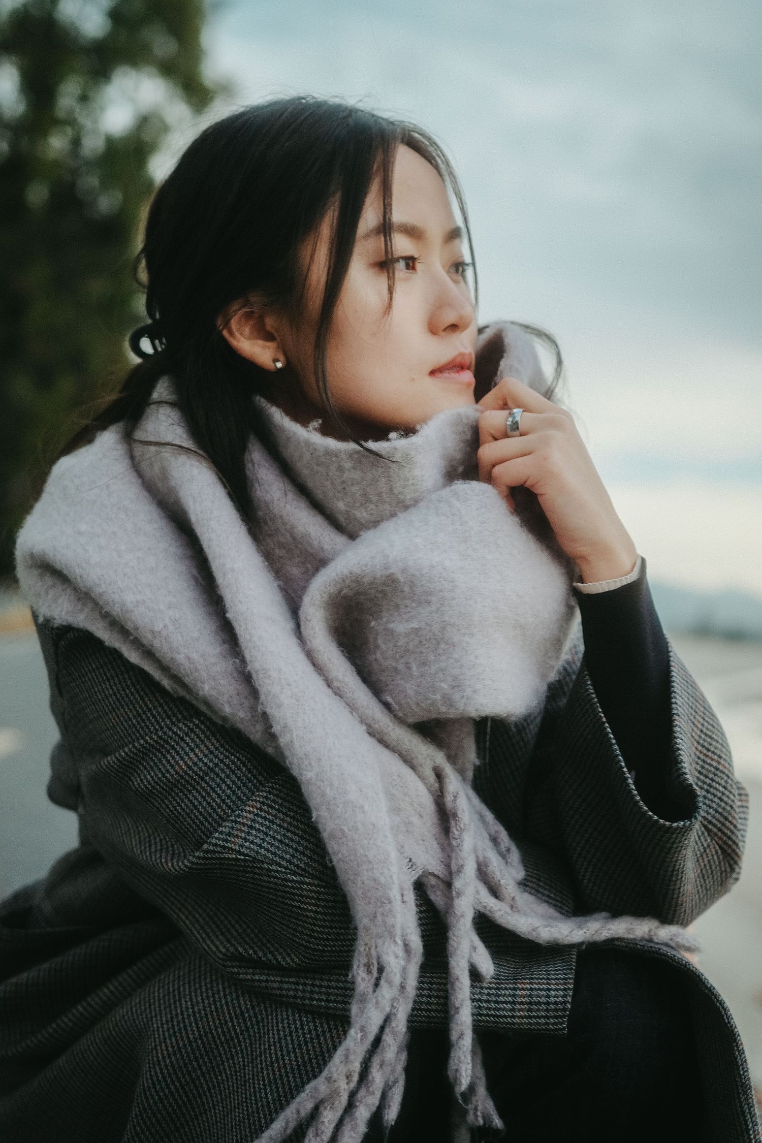 Young Asian woman in a gray plaid coat and soft scarf gazes thoughtfully outdoors on a cloudy day.