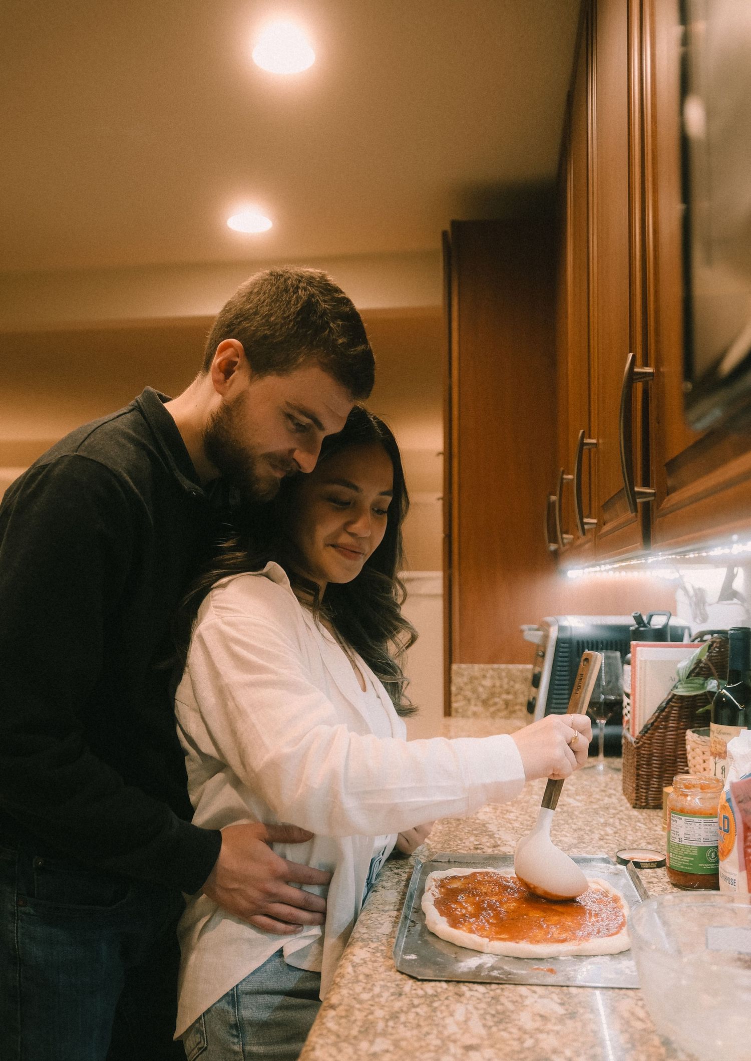 Couple making homemade pizza together in a warm kitchen, man embracing woman as she spreads tomato sauce on dough.