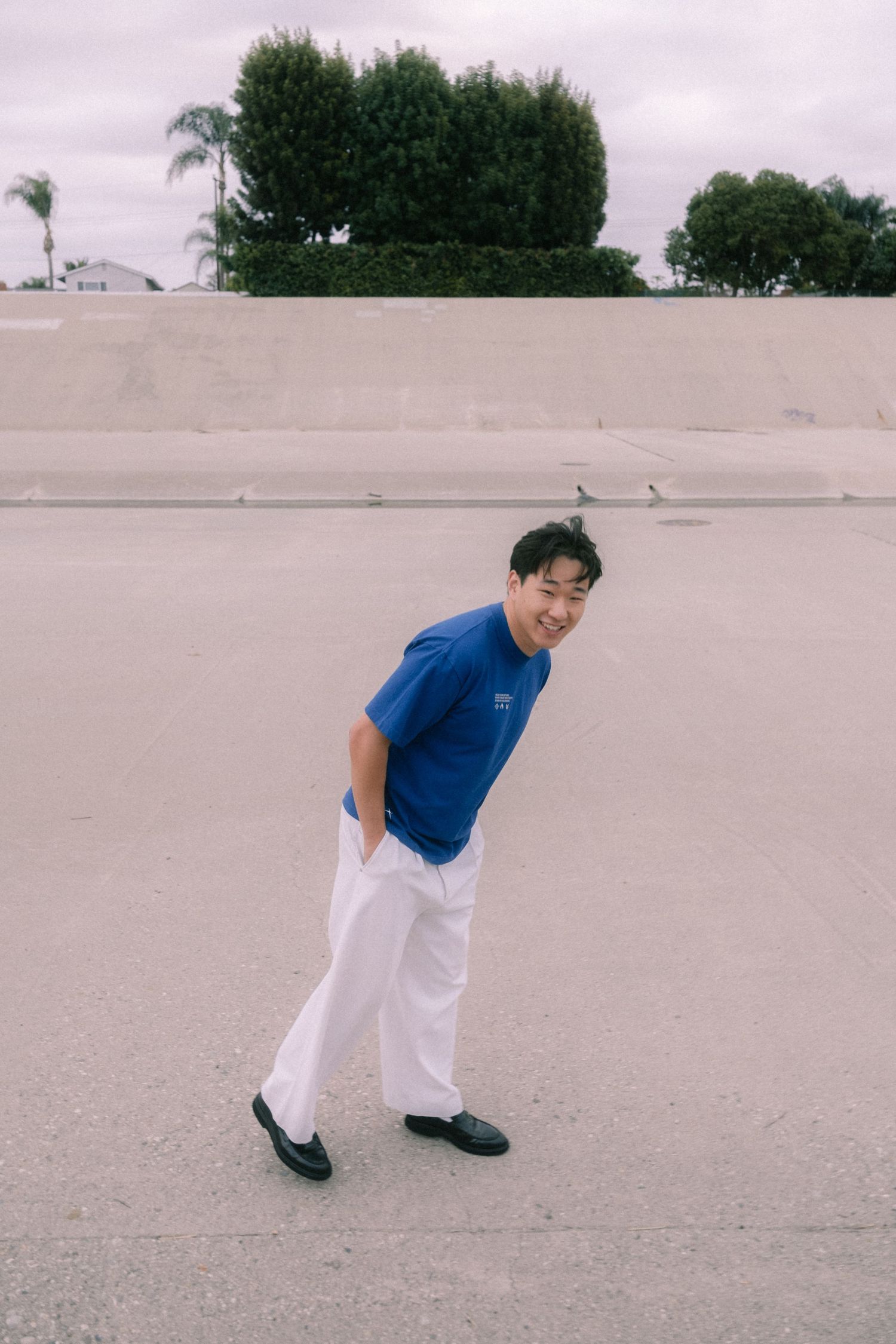 Smiling young man in blue tee and white pants posing on concrete plaza outdoors.
