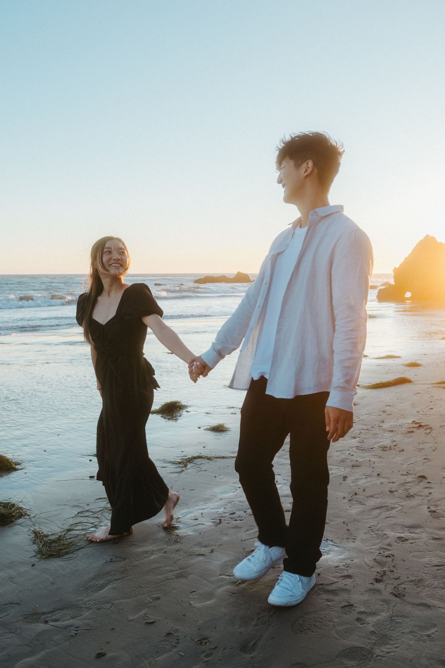 Happy couple holding hands and walking along a beach at sunset with rocky coastline in the background.