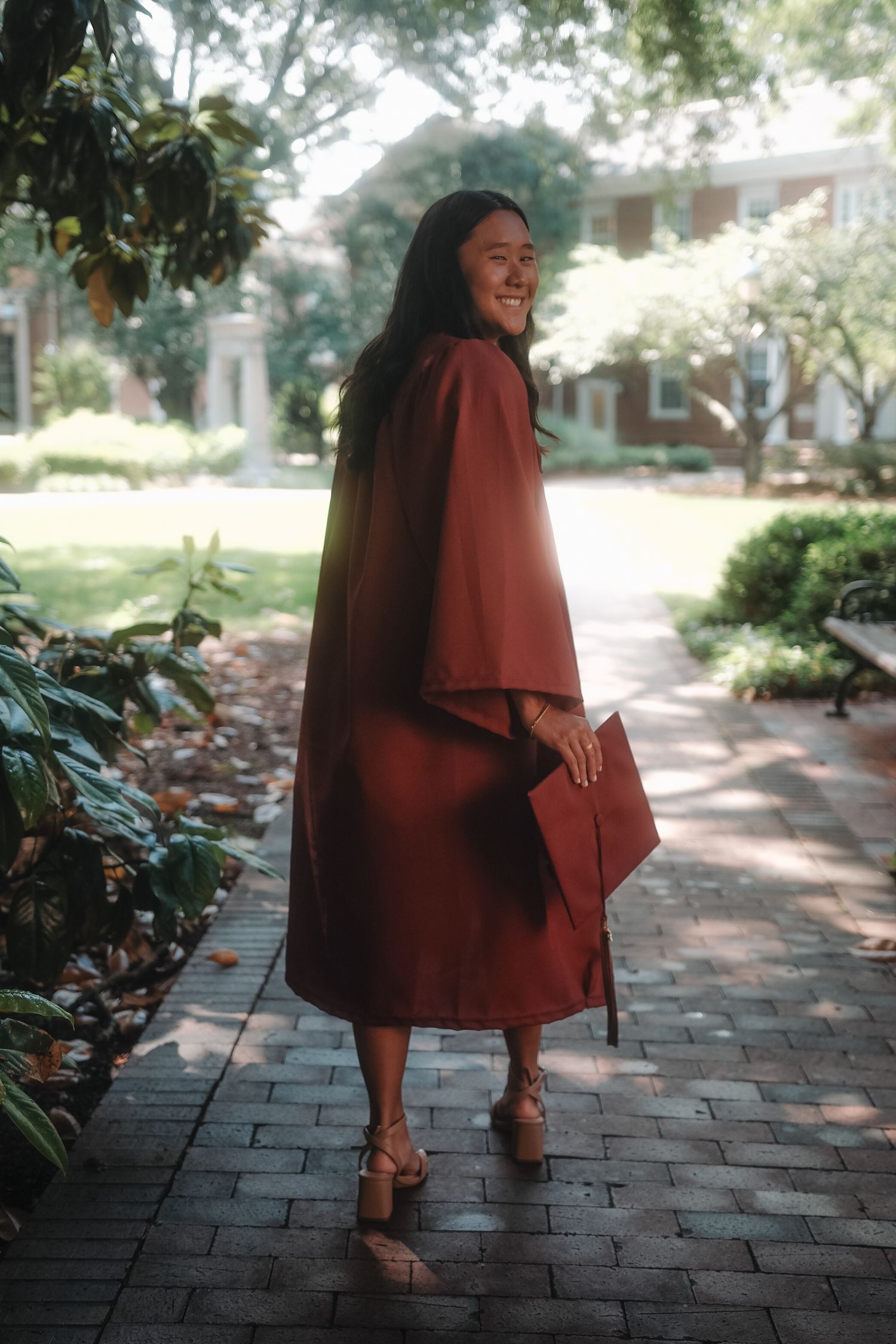 Graduate in maroon cap and gown walks along brick pathway looking back and smiling on campus.