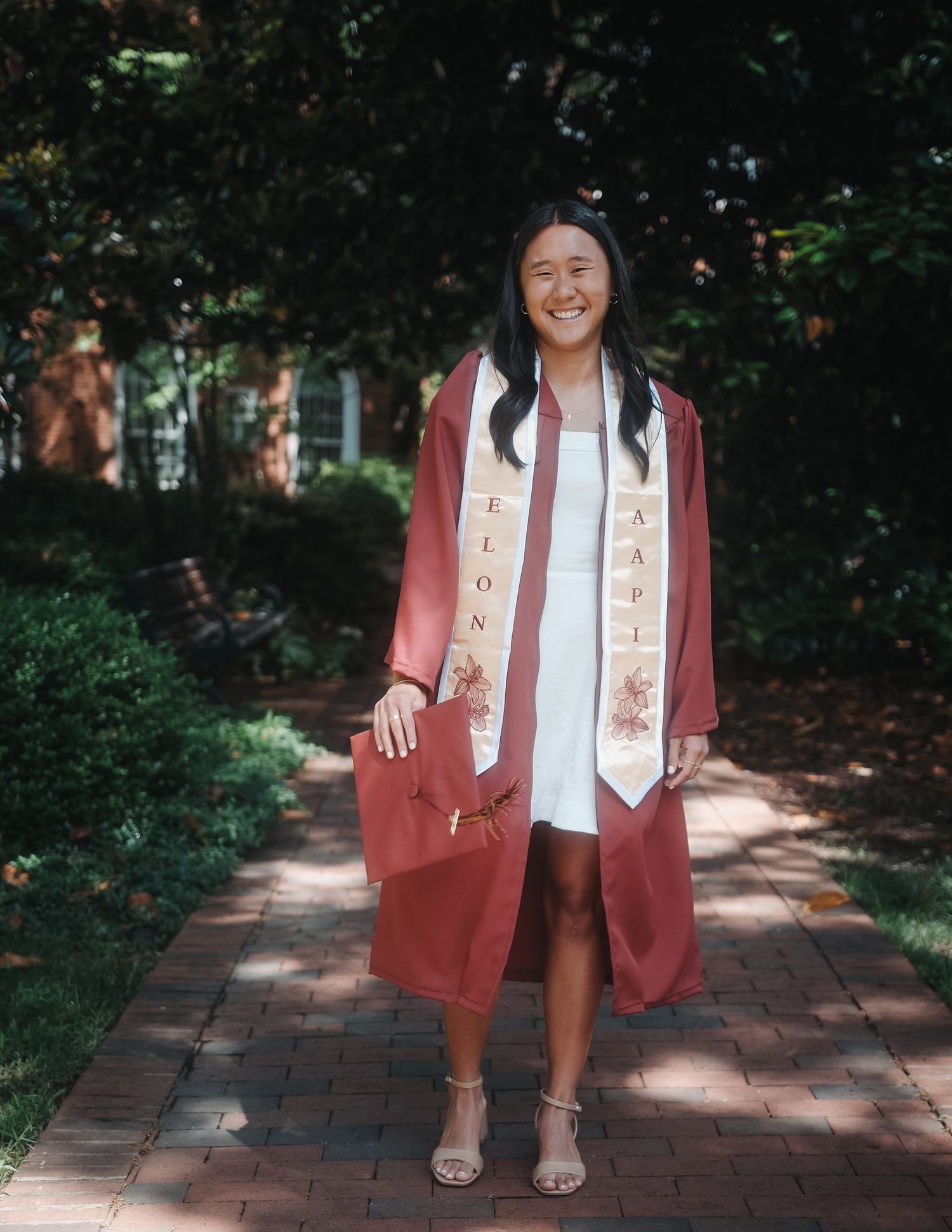 Smiling Elon graduate in maroon gown and honor stole holding cap on brick tree-lined walkway.