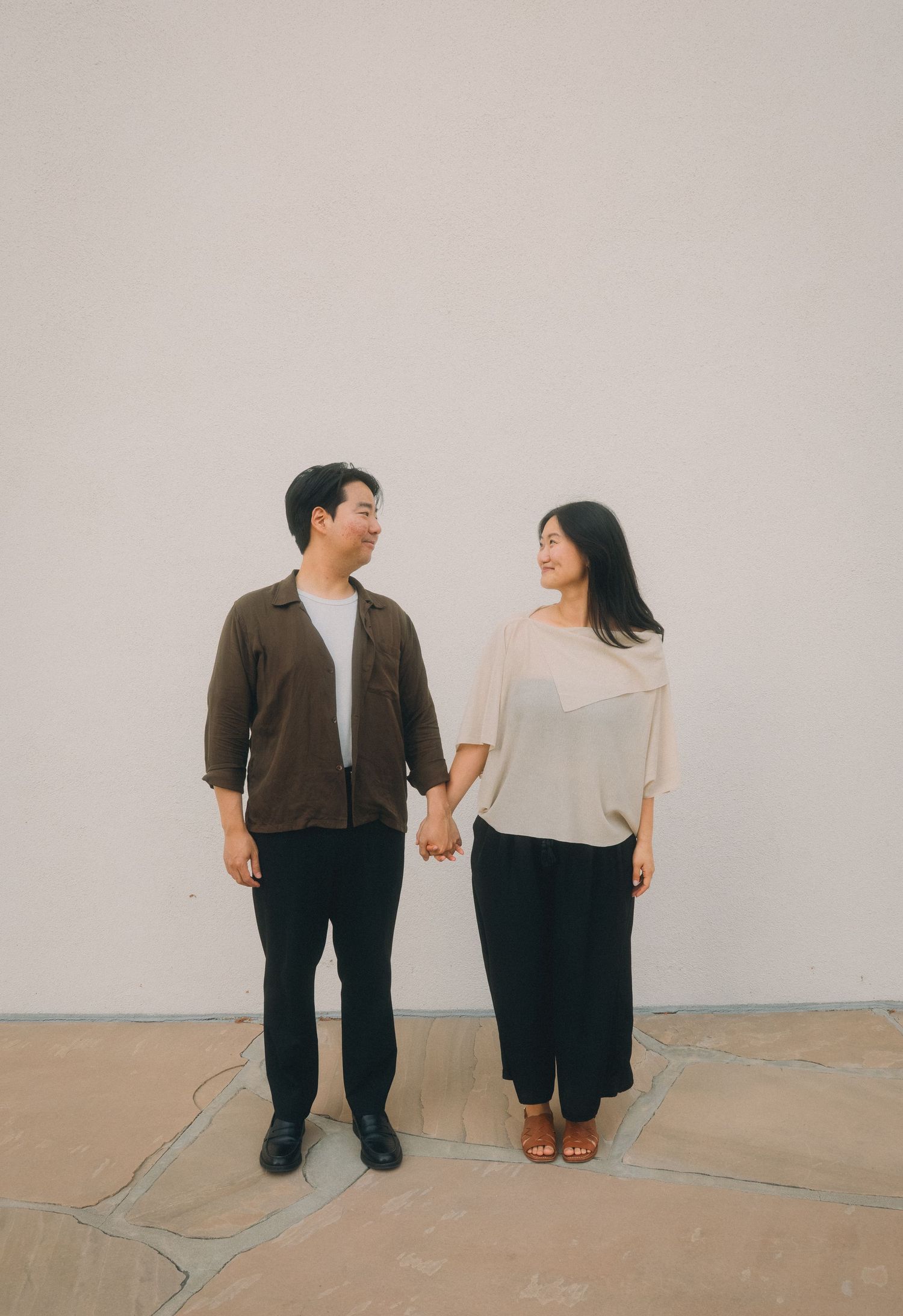 Asian couple holding hands and looking at each other lovingly against a white wall outdoors.