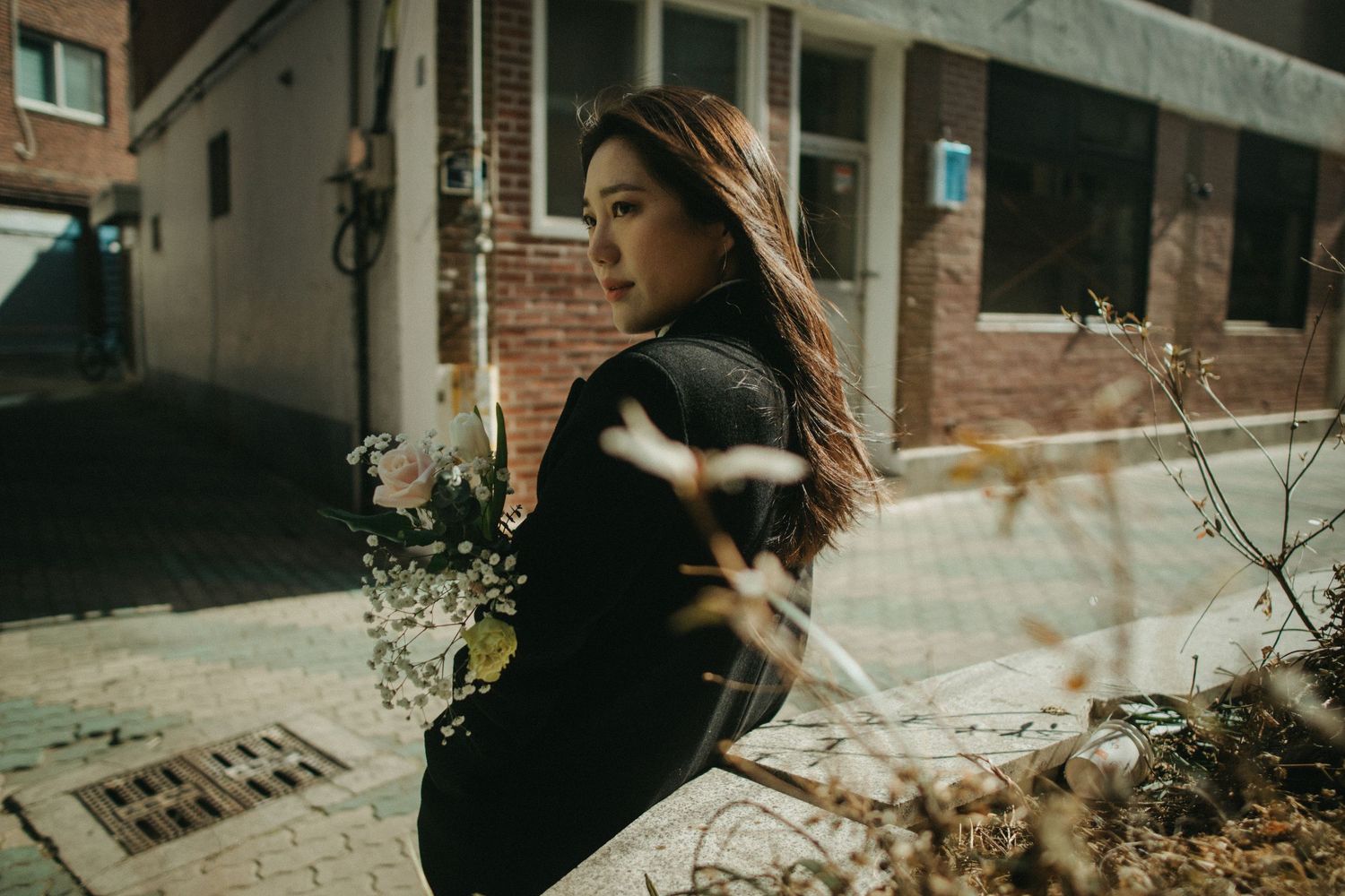 Young woman in black jacket holding bouquet, looking away on urban street in sunlight.