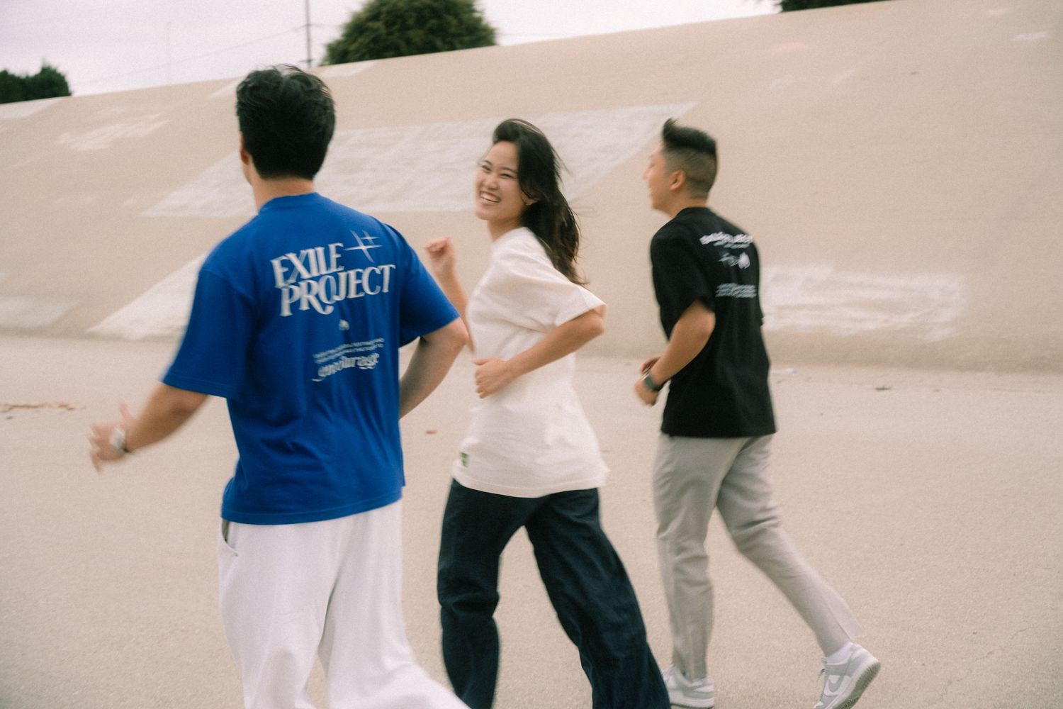 Three young people wearing casual streetwear walk and laugh together outdoors near a sandy wall.