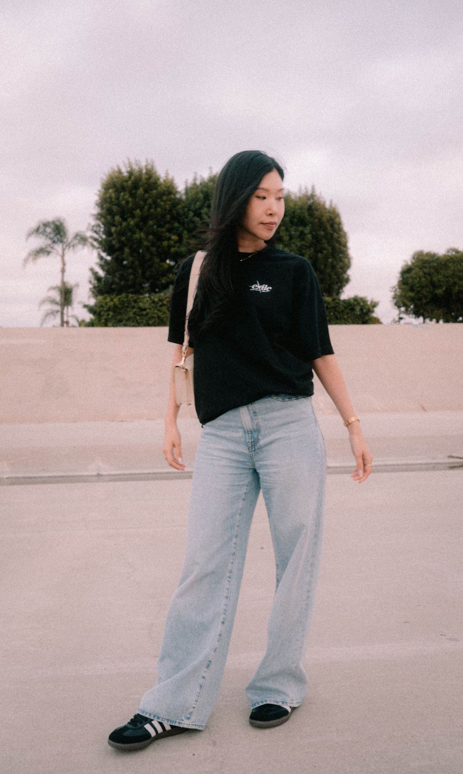 Young Asian woman in black t-shirt and wide-leg light wash jeans standing outdoors on overcast day.