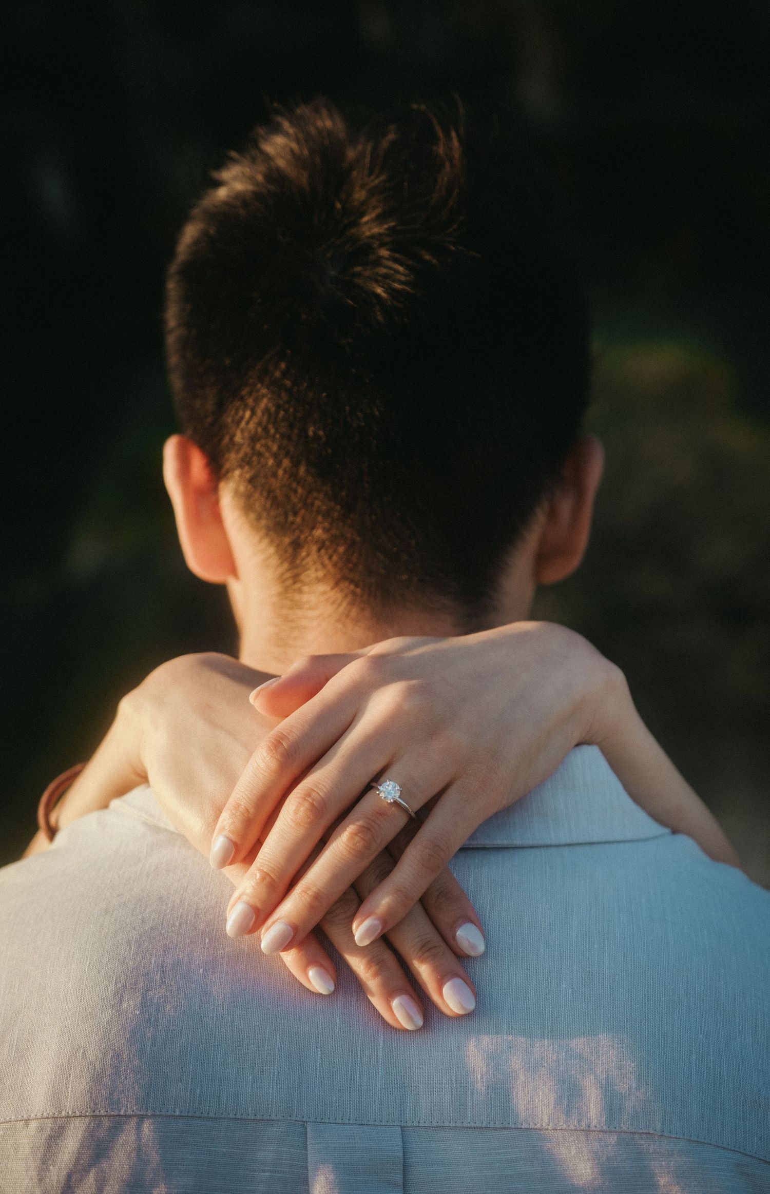 Close-up of a woman's hand with a diamond engagement ring resting on a man's shoulder.
