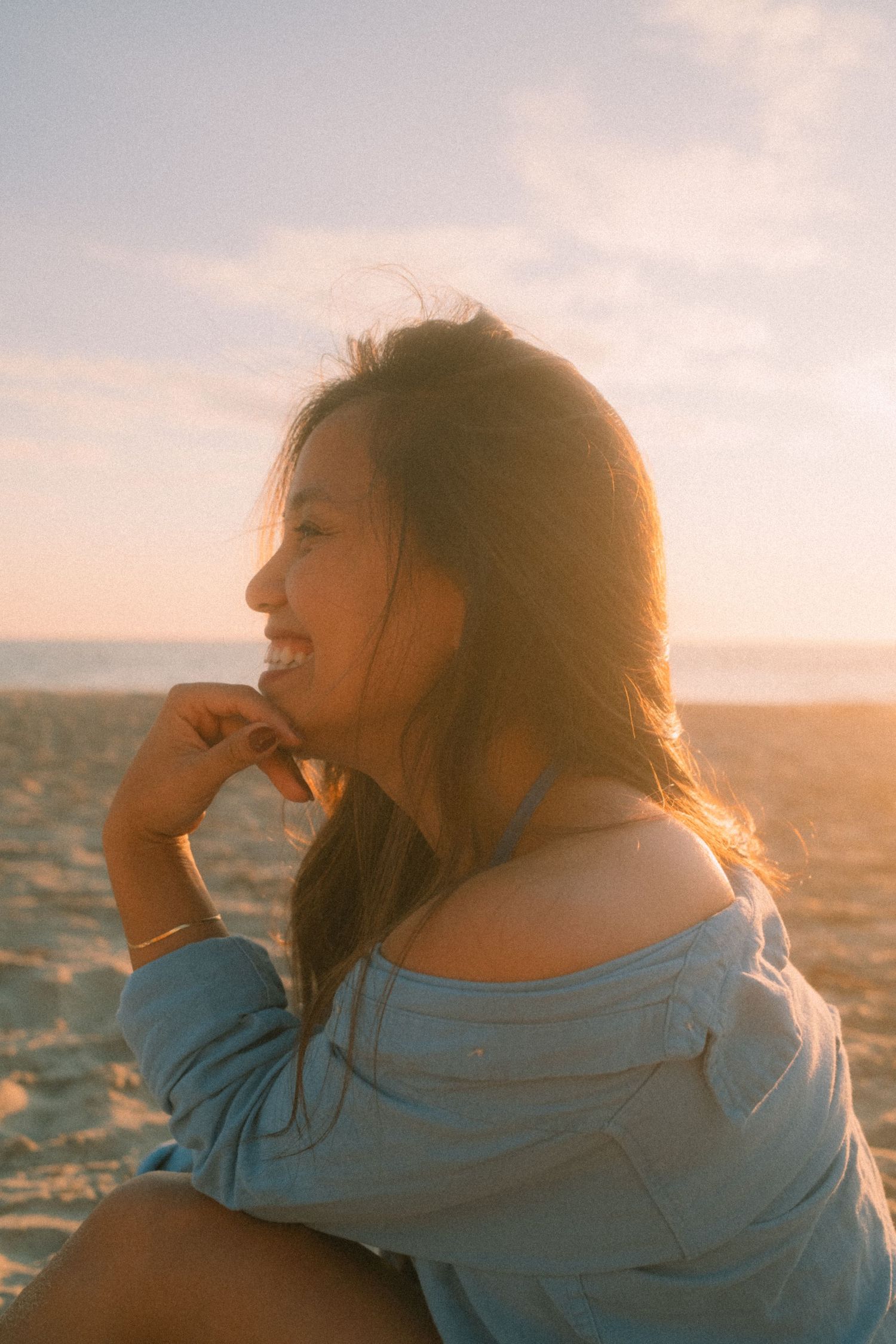 Cheerful young woman in blue off-shoulder top smiling on beach at sunset.