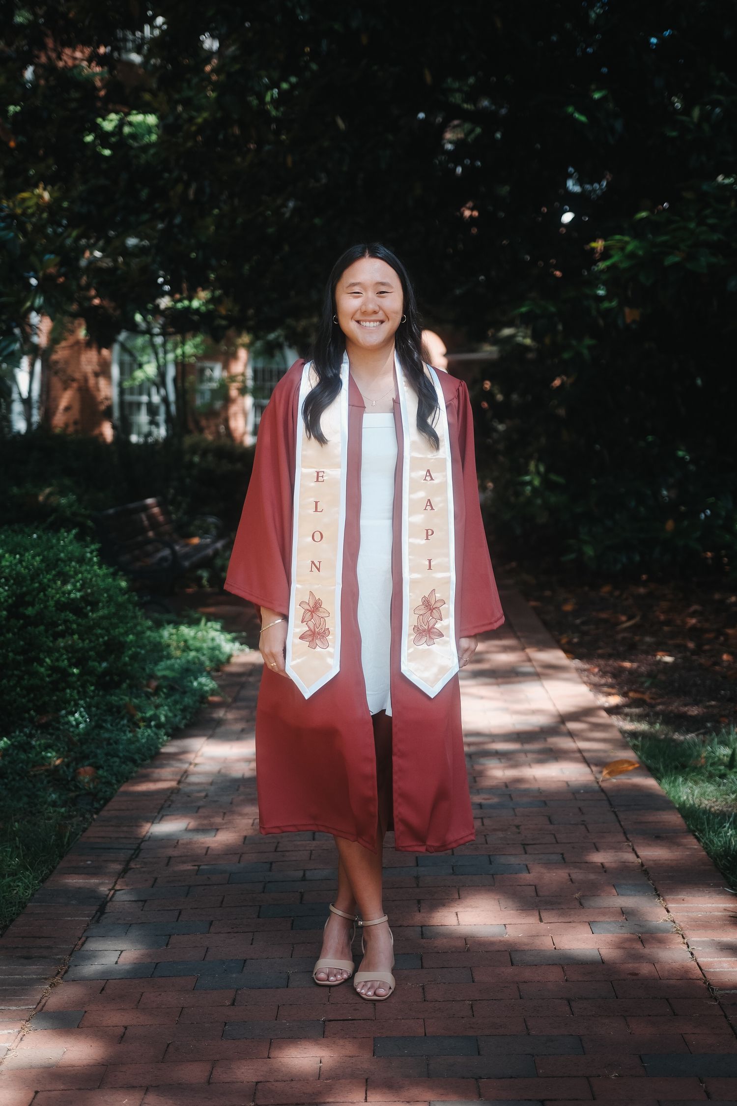Smiling graduate in maroon cap and gown with Elon University stole standing on brick path.