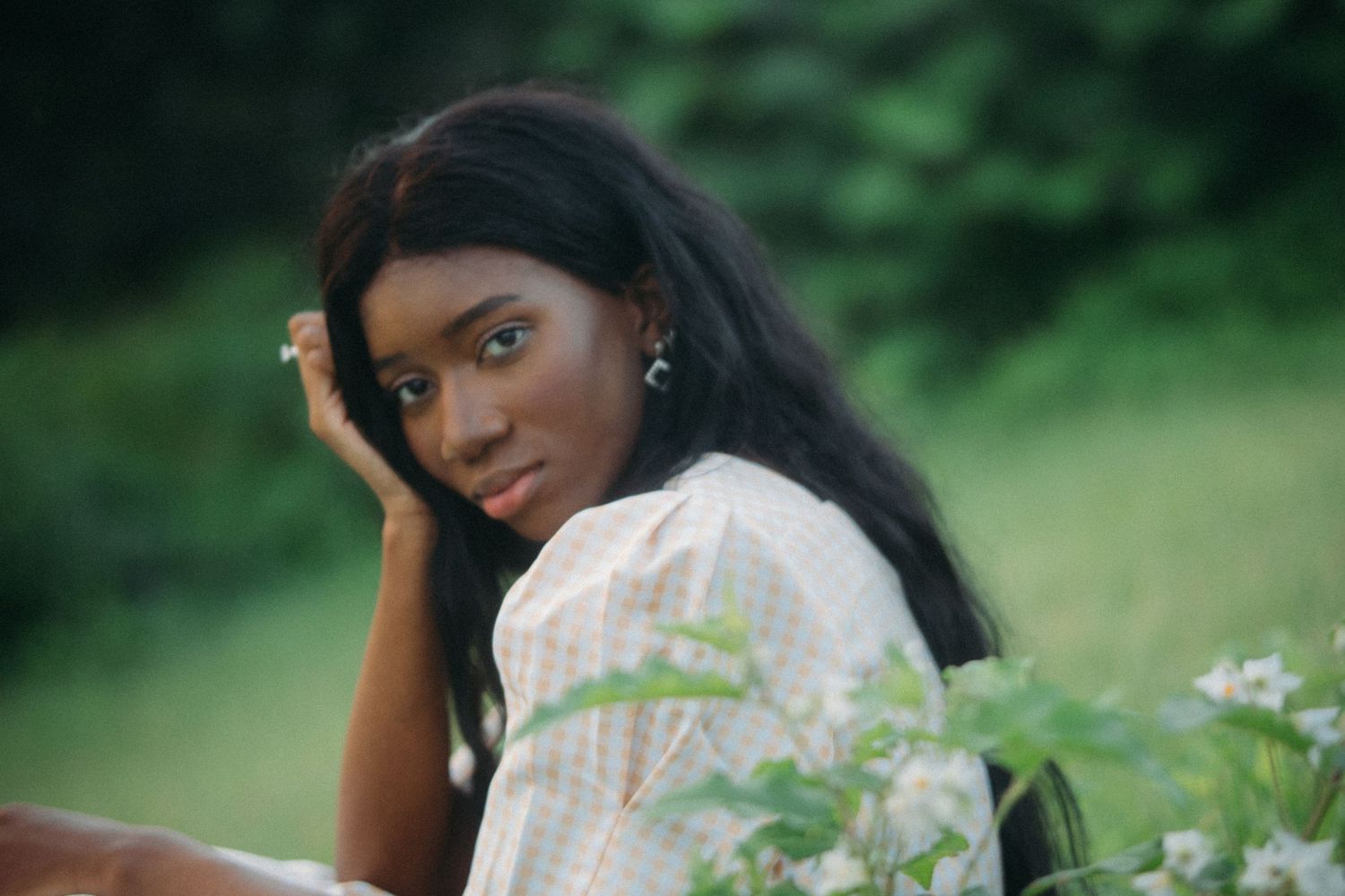 Young Black woman with long hair poses outdoors among green foliage and white flowers in a light floral dress.