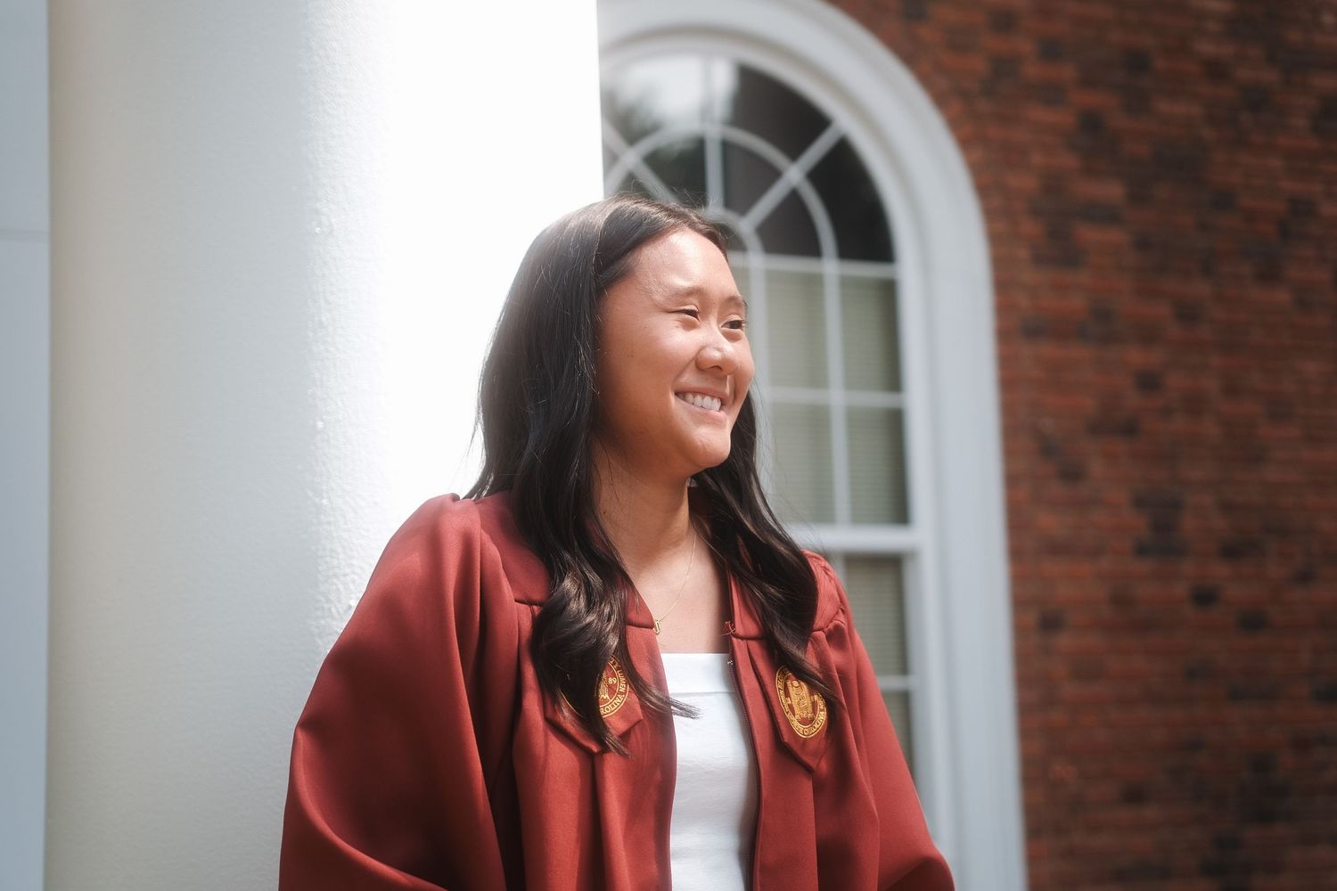 Joyful graduate in maroon gown laughs while standing near white column at Elon University campus.