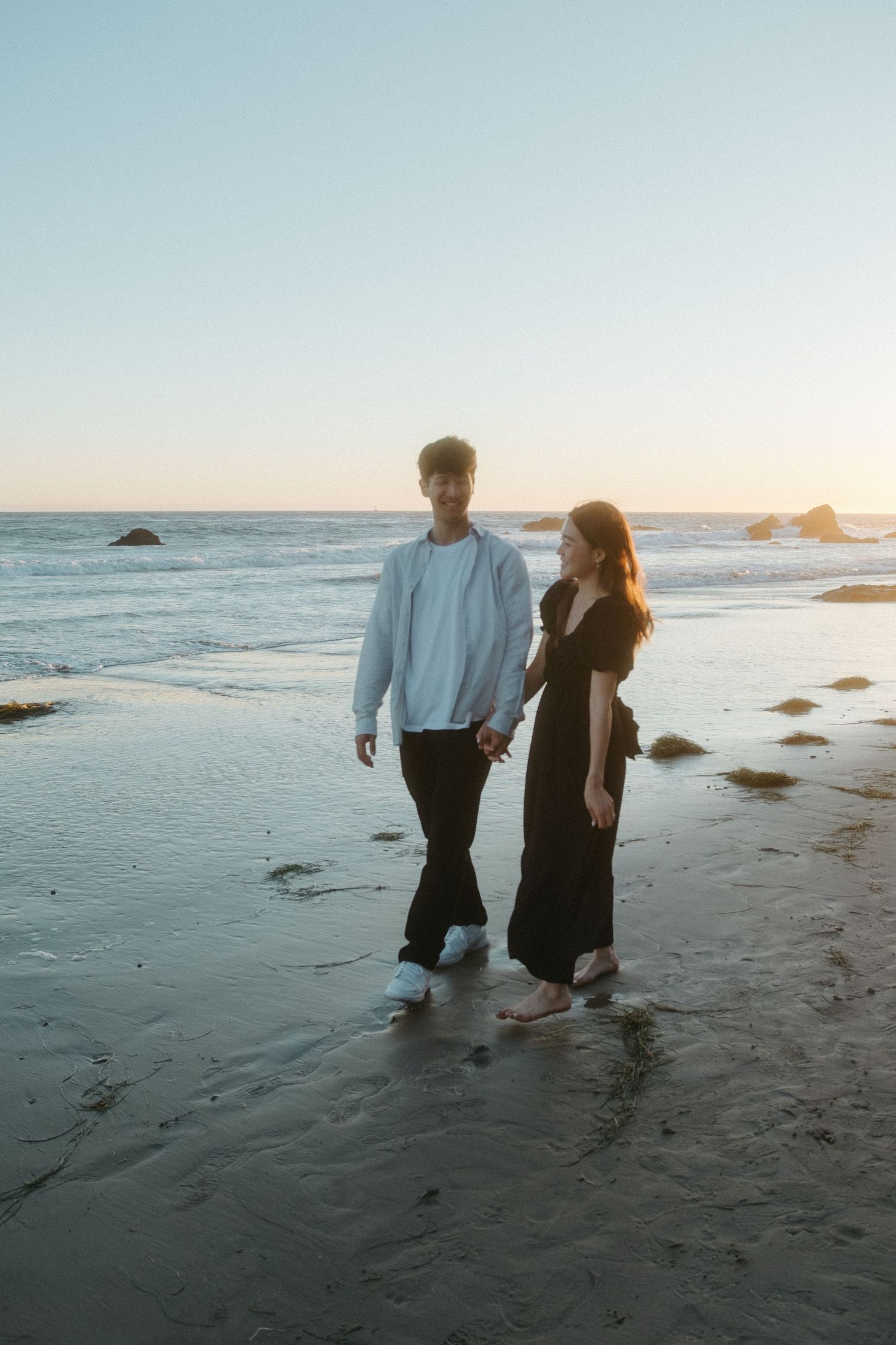 Couple walking hand in hand along a serene beach at sunset with ocean waves in the background.