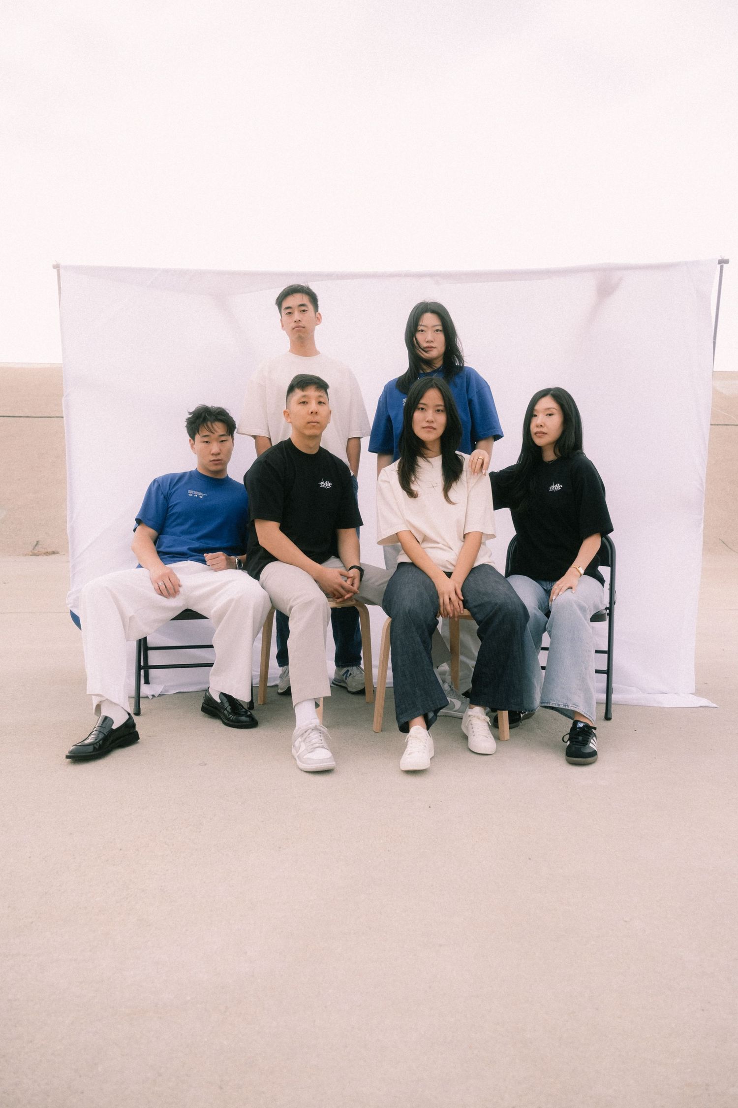 Group of six young adults posing in casual streetwear against a white backdrop outdoors.