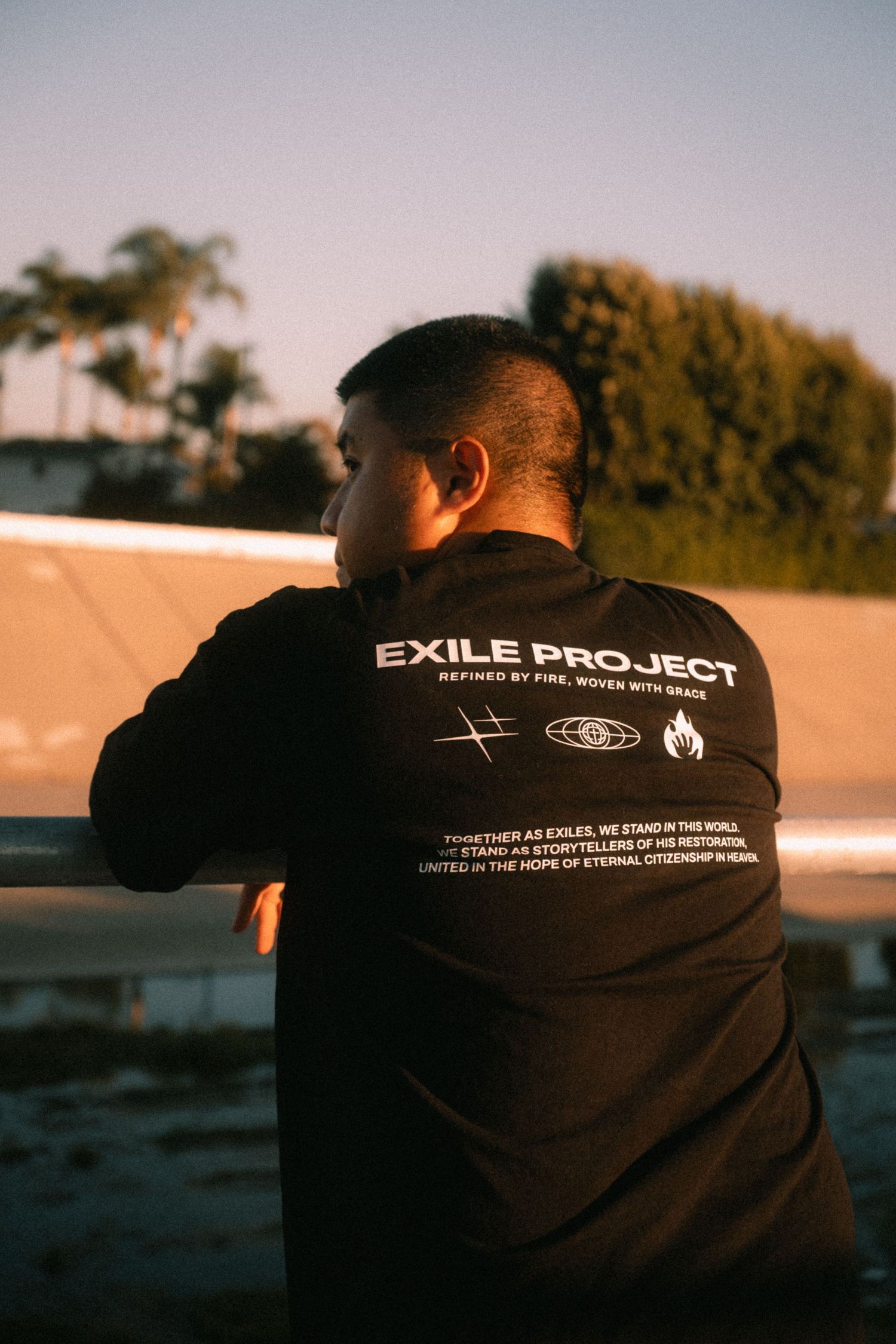 Man wearing black Exile Project hoodie with logo and text, photographed from behind at sunset.