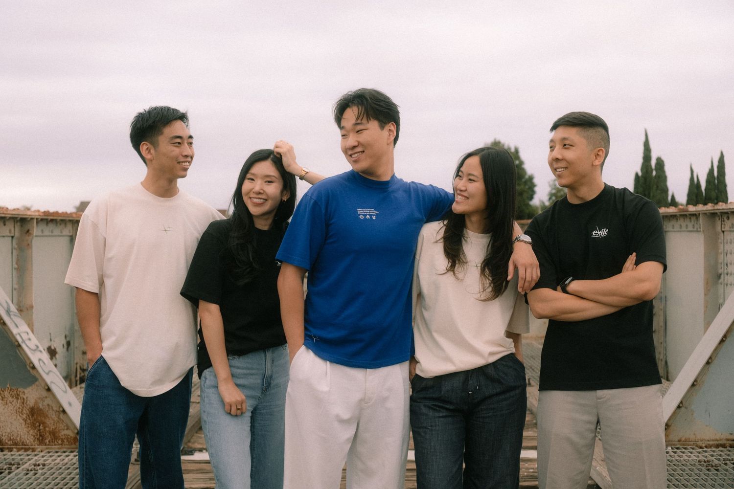 Group of five young Asian adults smiling together outdoors wearing casual t-shirts in neutral tones.