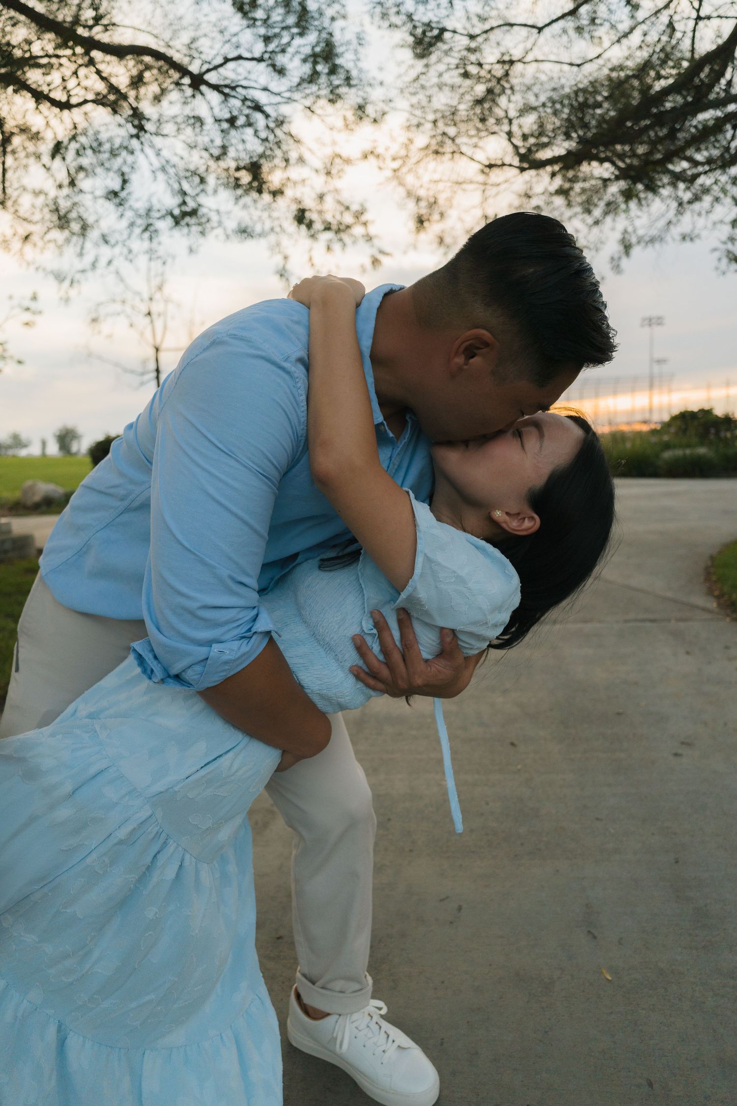 Couple sharing a romantic dip kiss outdoors at sunset, both wearing light blue outfits near a tree.