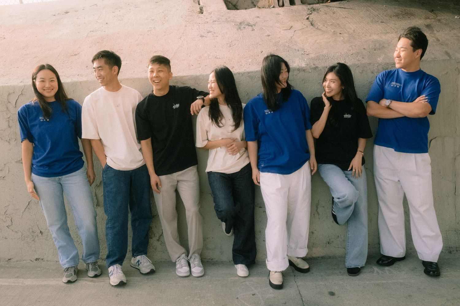 Seven young Asian adults in casual blue and white outfits laughing together outdoors against a sandy wall.