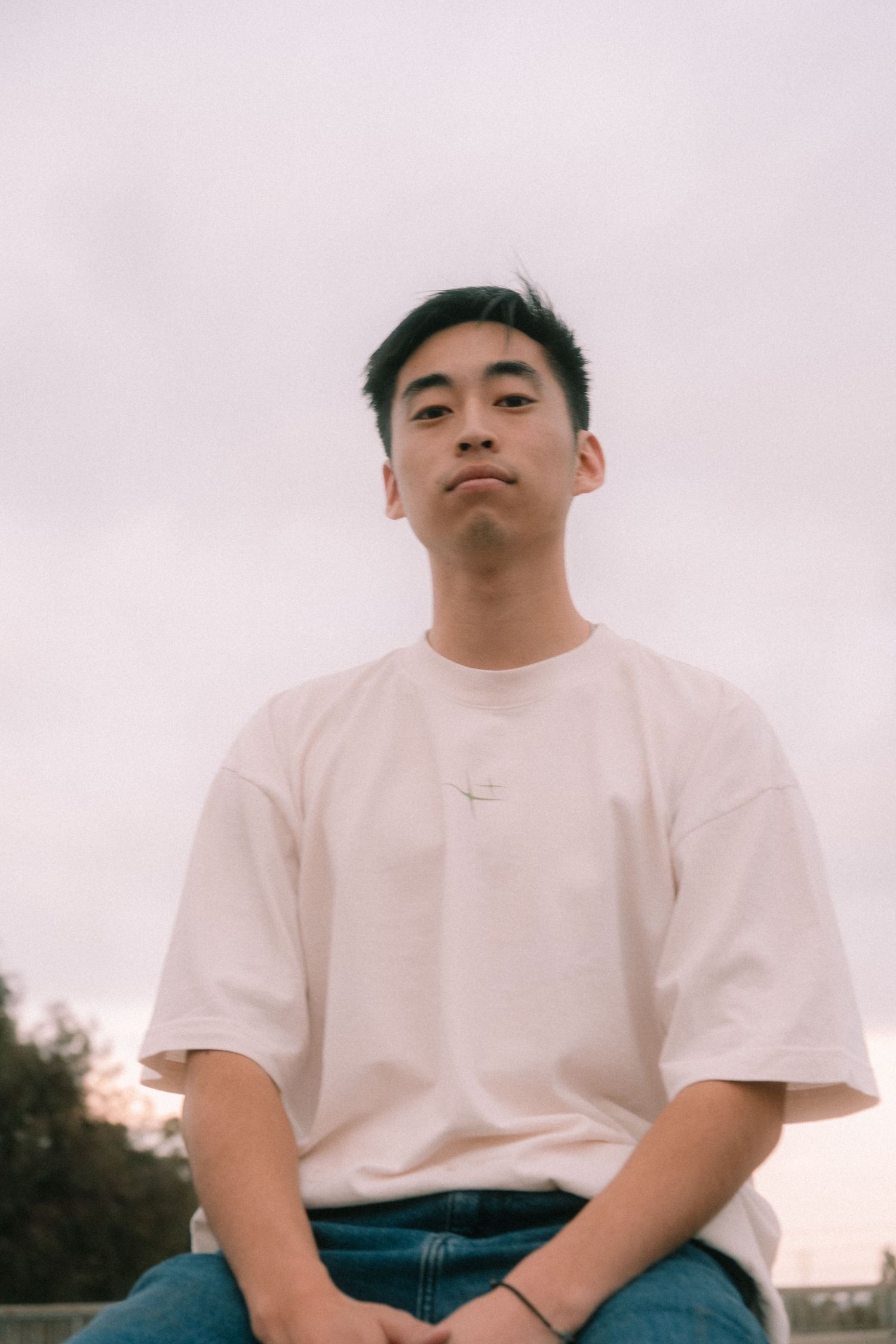 Young Asian man in white oversized t-shirt sitting casually against an overcast sky background.