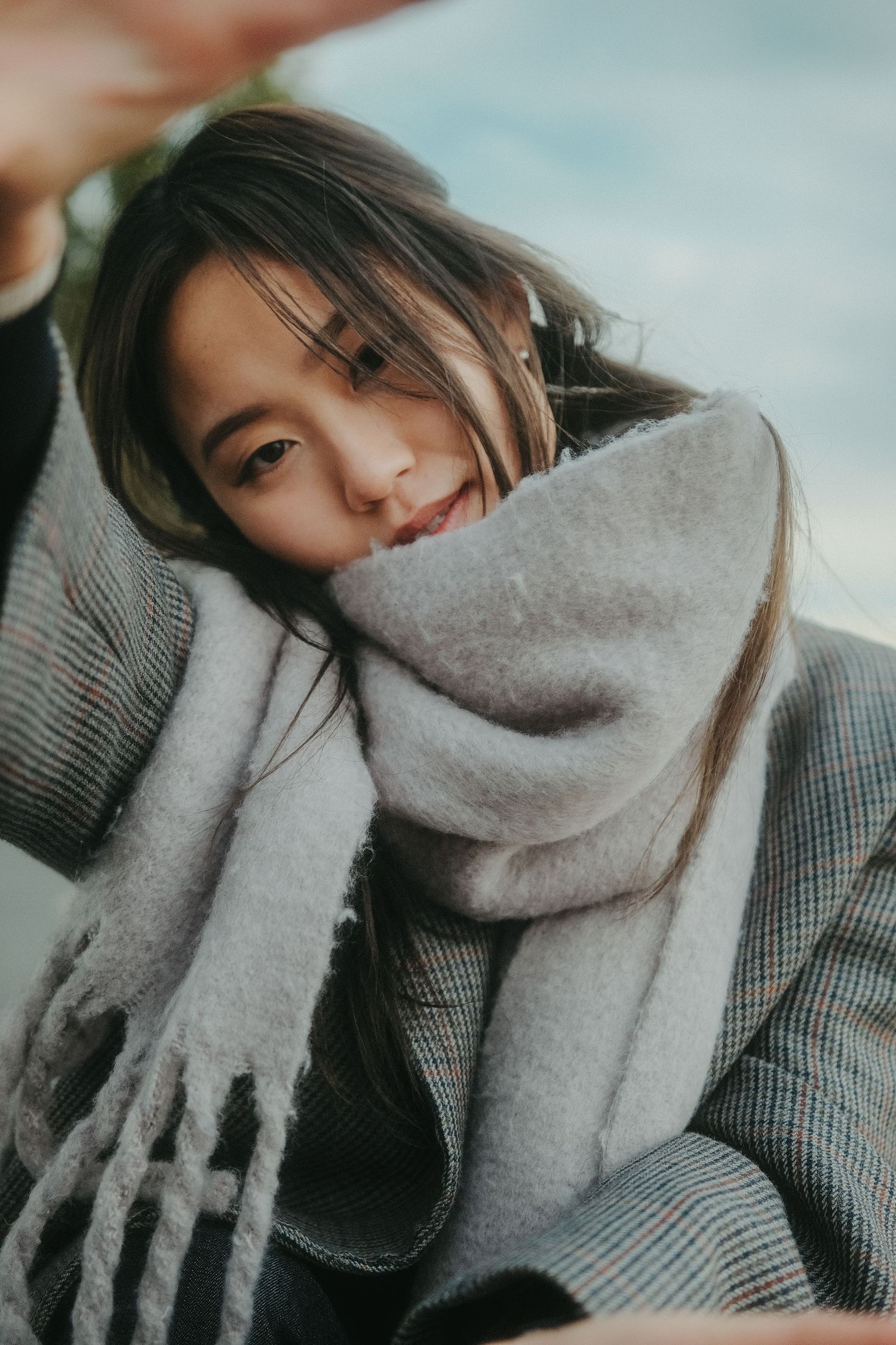 Young Asian woman wrapped in a cozy gray scarf and plaid coat, looking up at the camera outdoors.