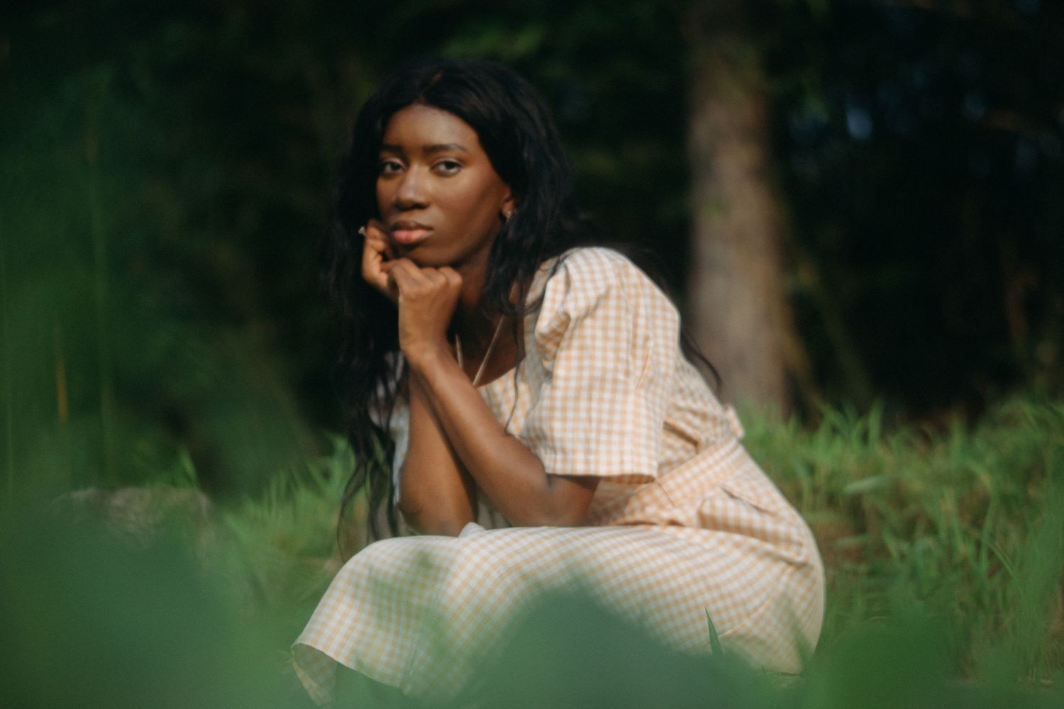 Young Black woman in checkered dress sits thoughtfully in lush green nature setting, hands clasped under chin.